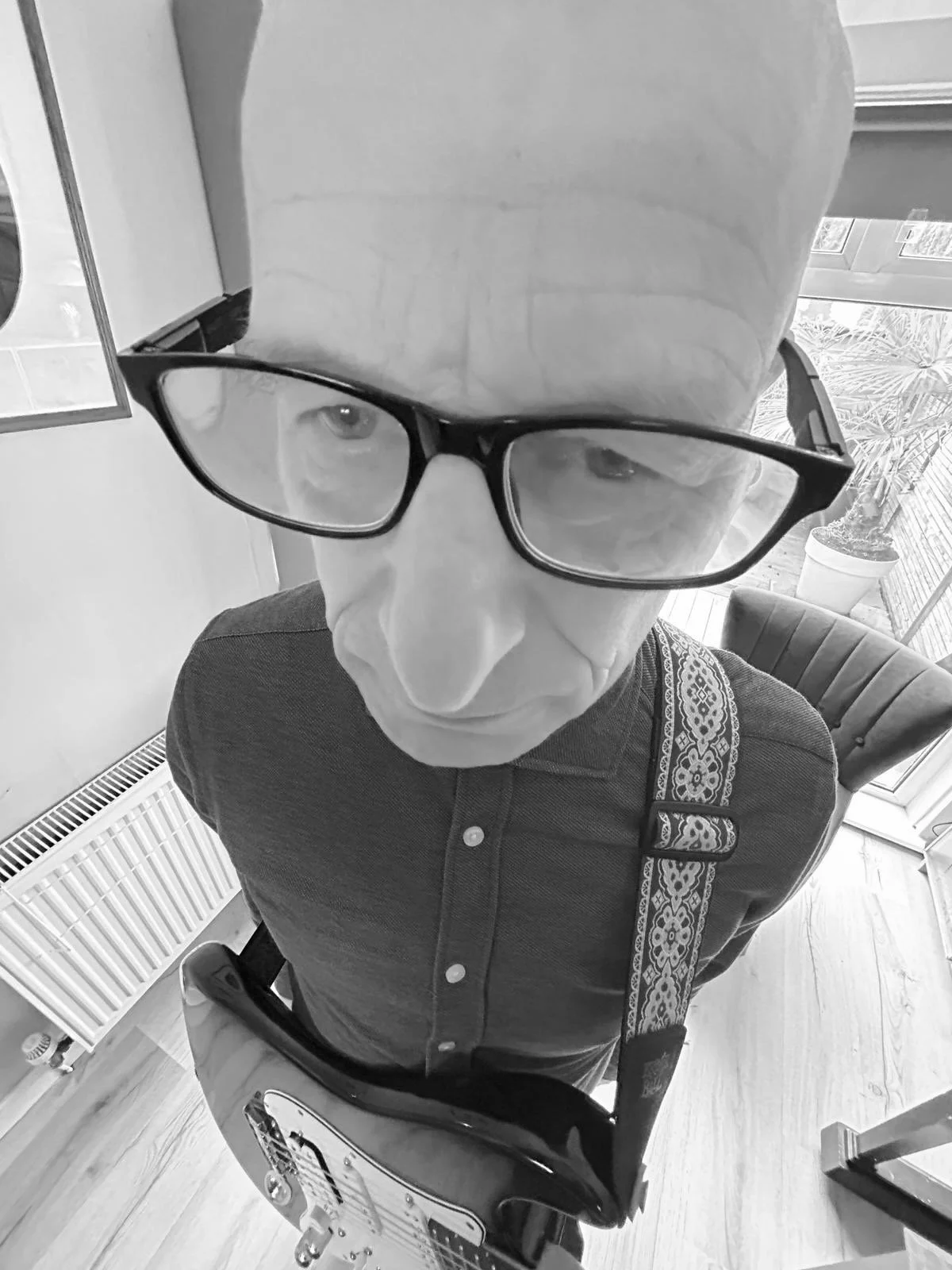 A black-and-white selfie taken from above shows a man with glasses, a bald head, and light skin. He is wearing a dark button-up shirt and carrying a guitar strap. He is in a room with wooden flooring, a plant in a pot, and a window with blinds in the background.