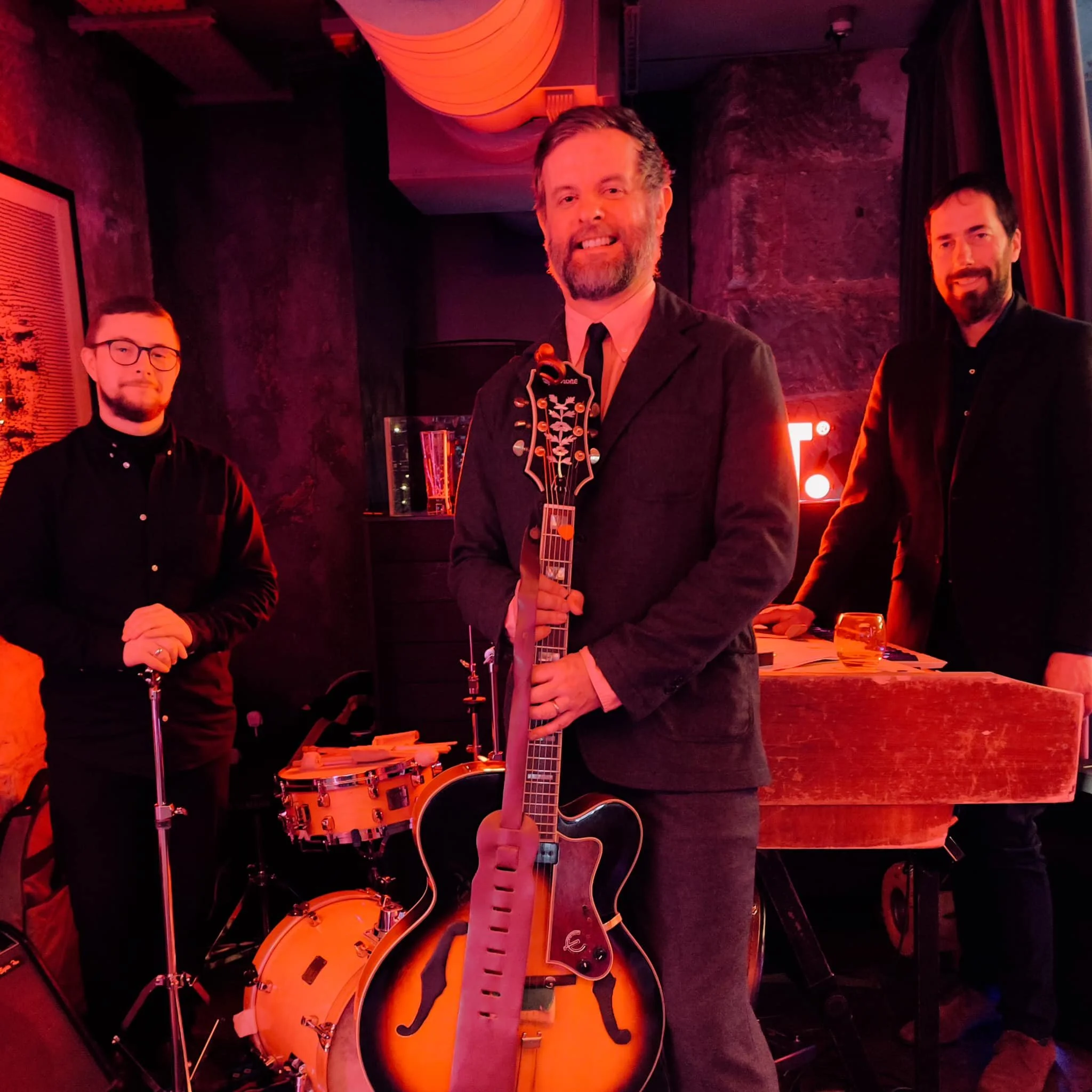 A group of men at a social event, standing indoors with low red lighting, some holding drinks, with musical instruments and decorations in the background.