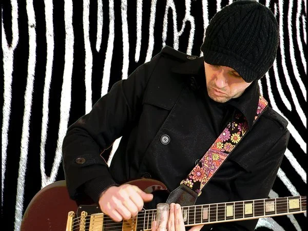 Man wearing black knit cap and black jacket playing an electric guitar with a floral strap, against a black and white striped background.
