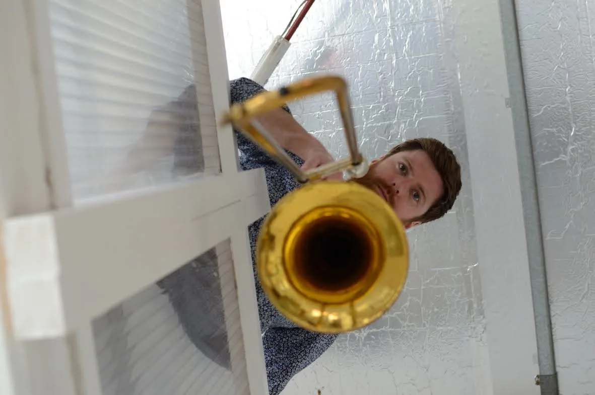 A man is looking up at a camera while playing a trumpet, with the photo taken from below through a glass window.