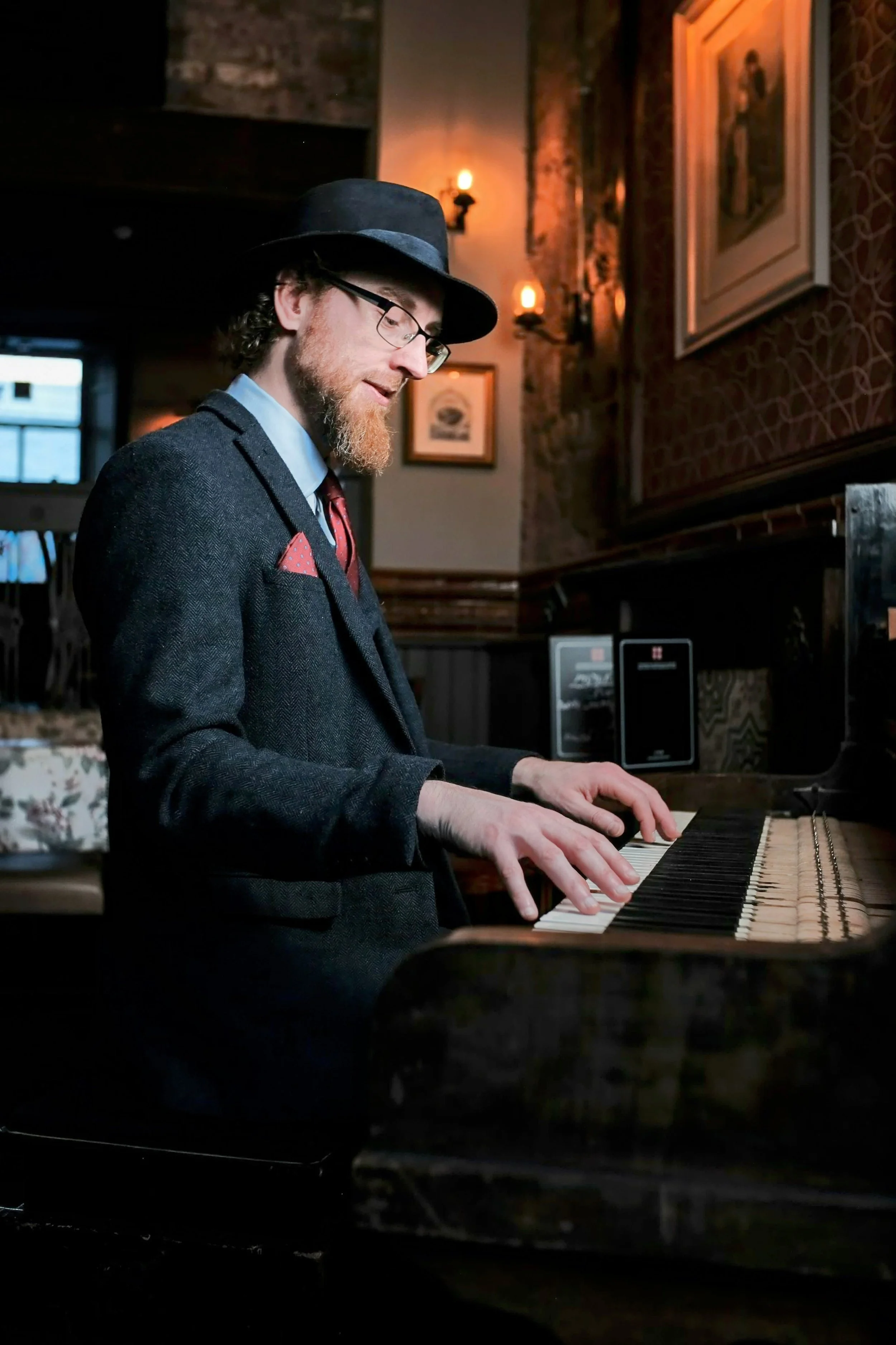 Man dressed in vintage suit playing a piano in a dimly lit, cozy room with artwork on the walls and warm lighting.