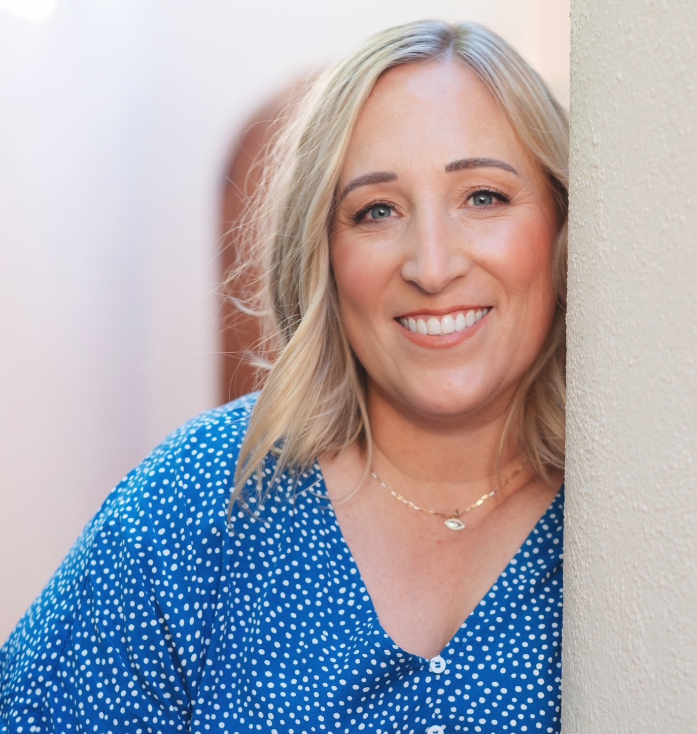 Person with blonde hair wearing a light-colored blouse, smiling at the camera against a plain background.