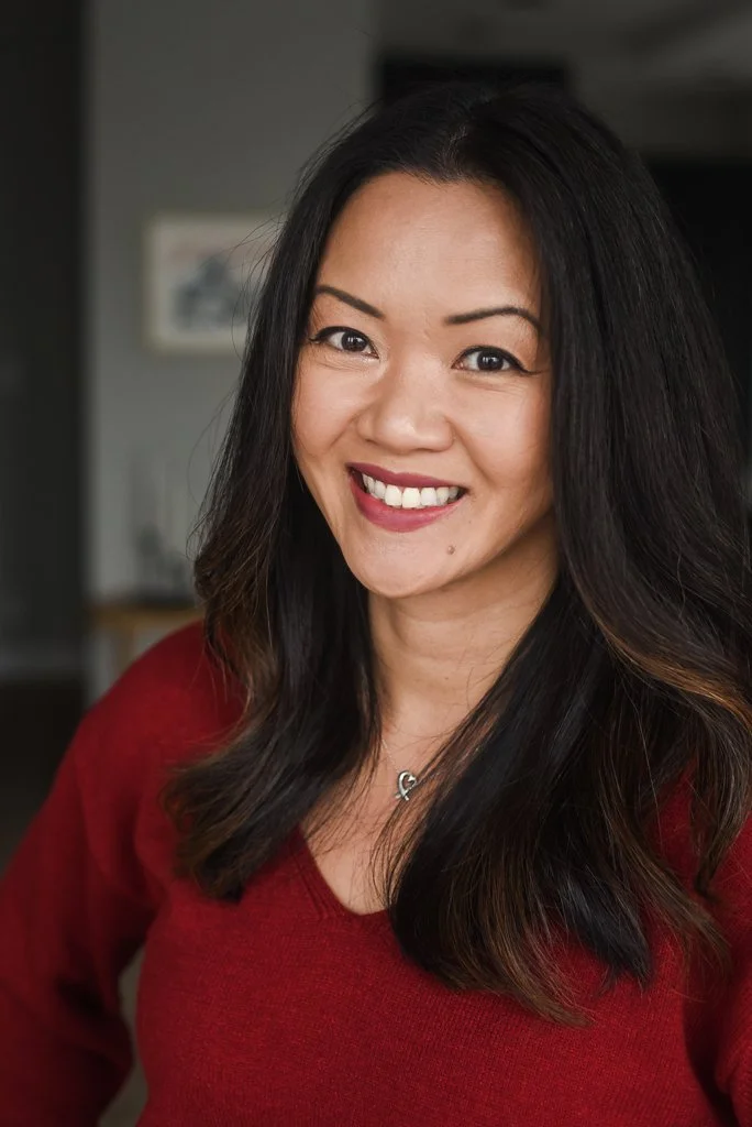 A woman with long dark hair, smiling, wearing a red top and a necklace with a heart-shaped pendant, in an indoor setting.