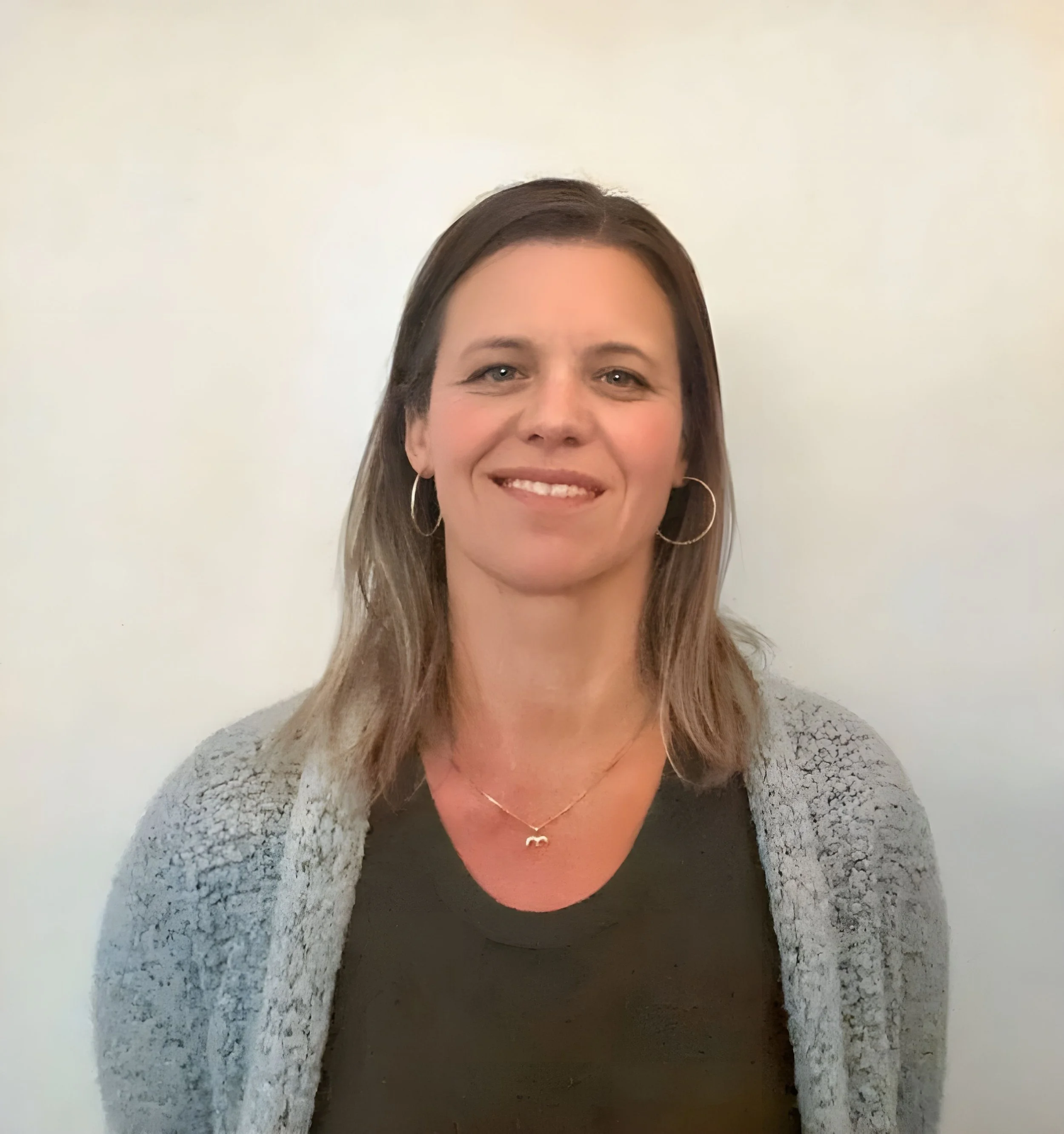 A woman with shoulder-length brown hair, wearing hoop earrings, a black top, a gray textured cardigan, and a necklace with a small infinity symbol, standing against a white wall.
