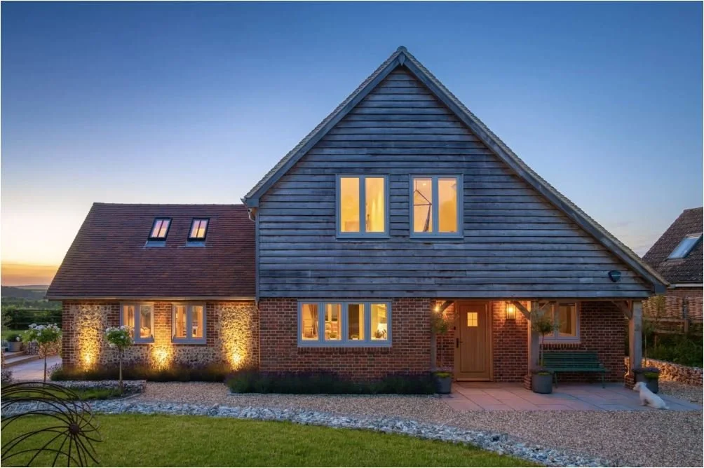 Gabled entrance elevation of Hill Top House at dusk, with timber cladding, brickwork and illuminated windows.