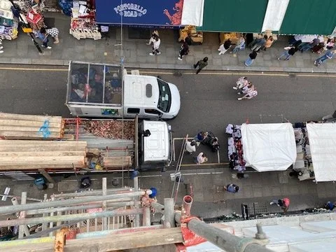 View from rooftop to ground - Scaffold lorry and tower being built with market traders and public surrounding the work site. Homebuilding Essentials
