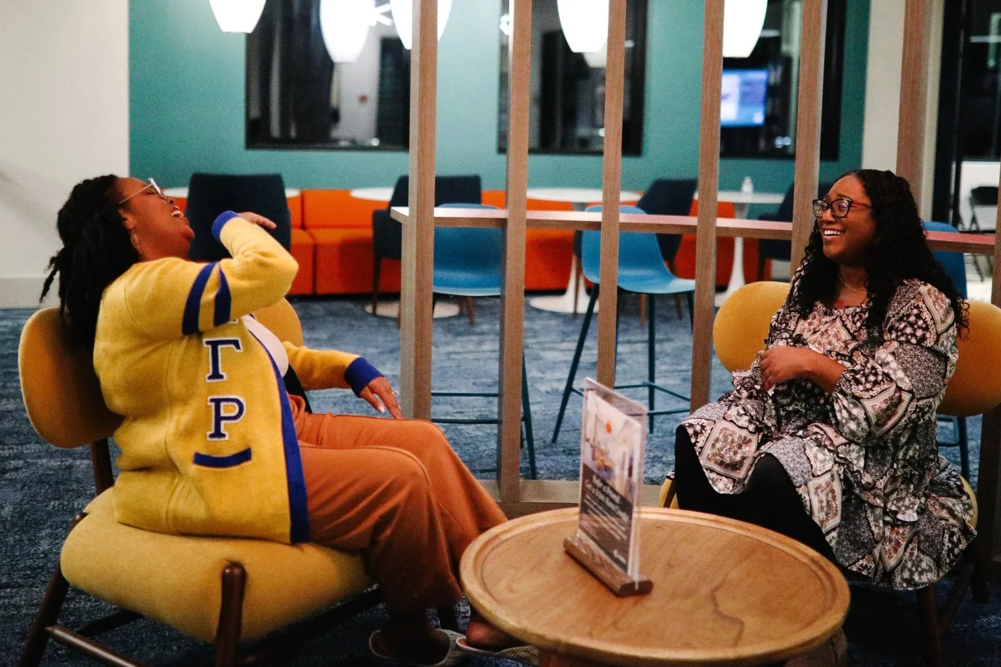 Two women sitting and laughing in a lounge area, one wearing a yellow jacket and the other in a patterned dress, separated by a wooden partition, with chairs and a television in the background.