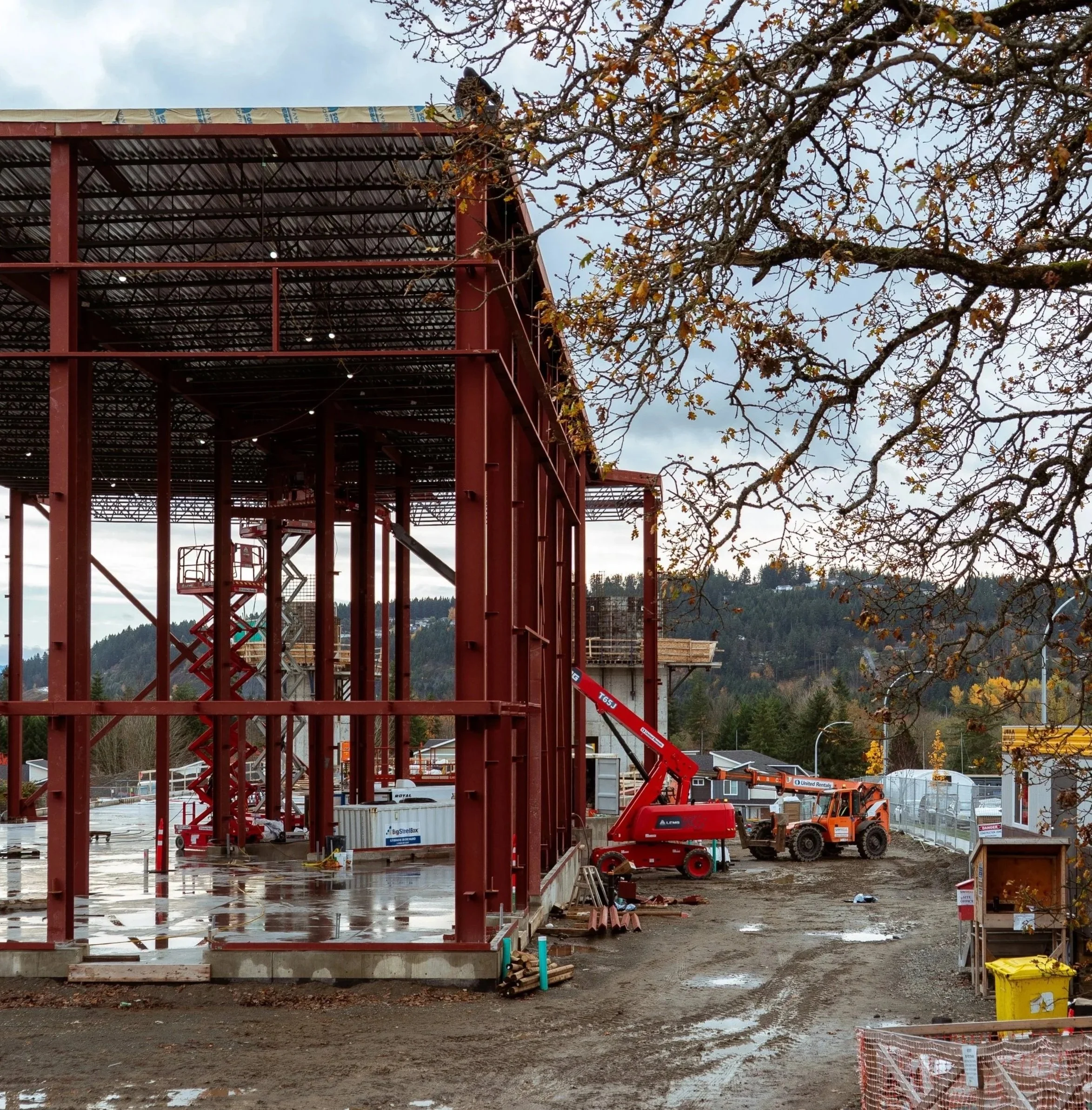 Under construction building with red steel framework, construction equipment, and machinery on a muddy site with puddles, surrounded by trees and a cloudy sky.
