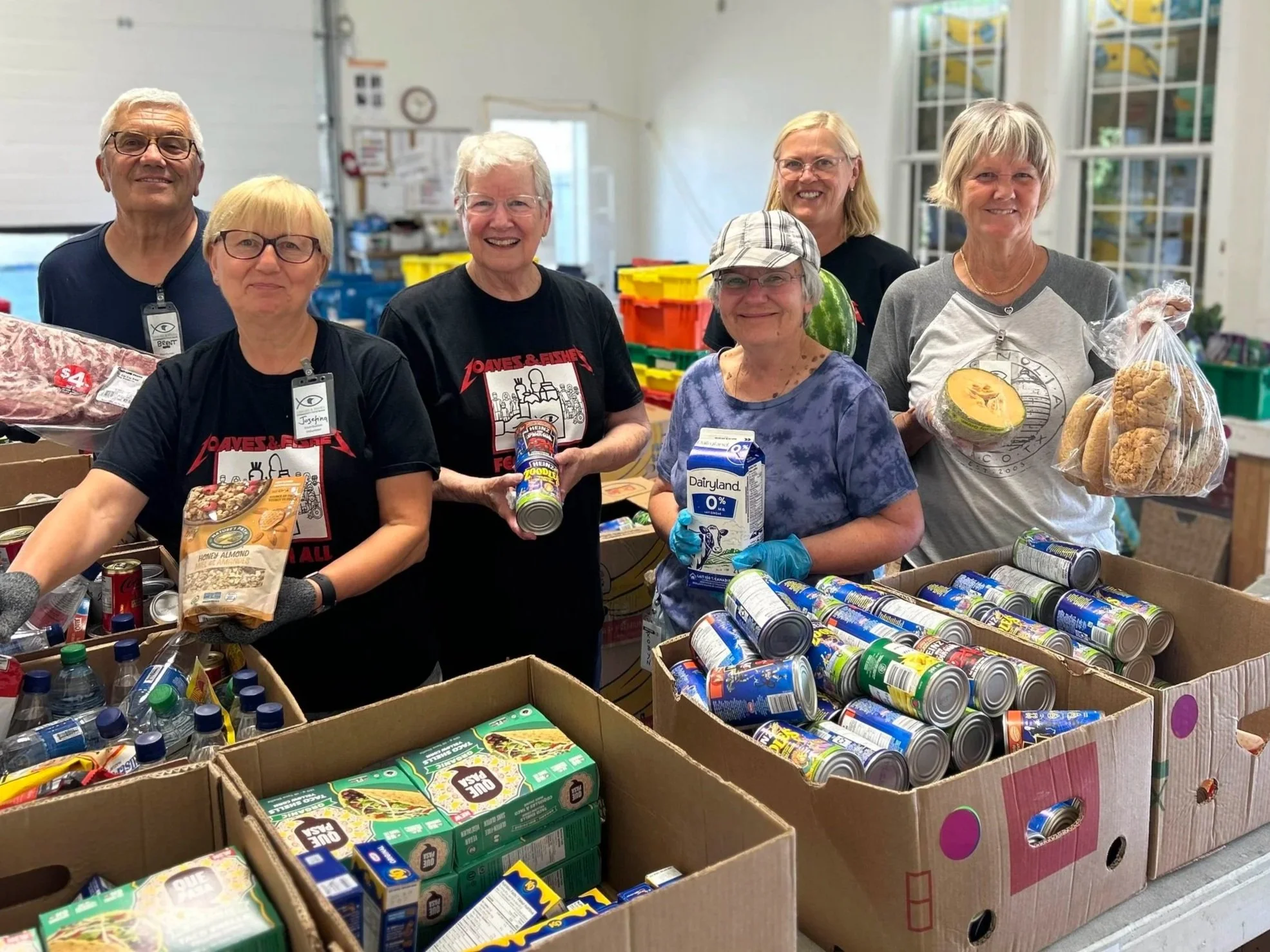 Group of seven volunteers packing groceries at a food bank or community pantry, with boxes of canned goods, snacks, and other food items on the table.