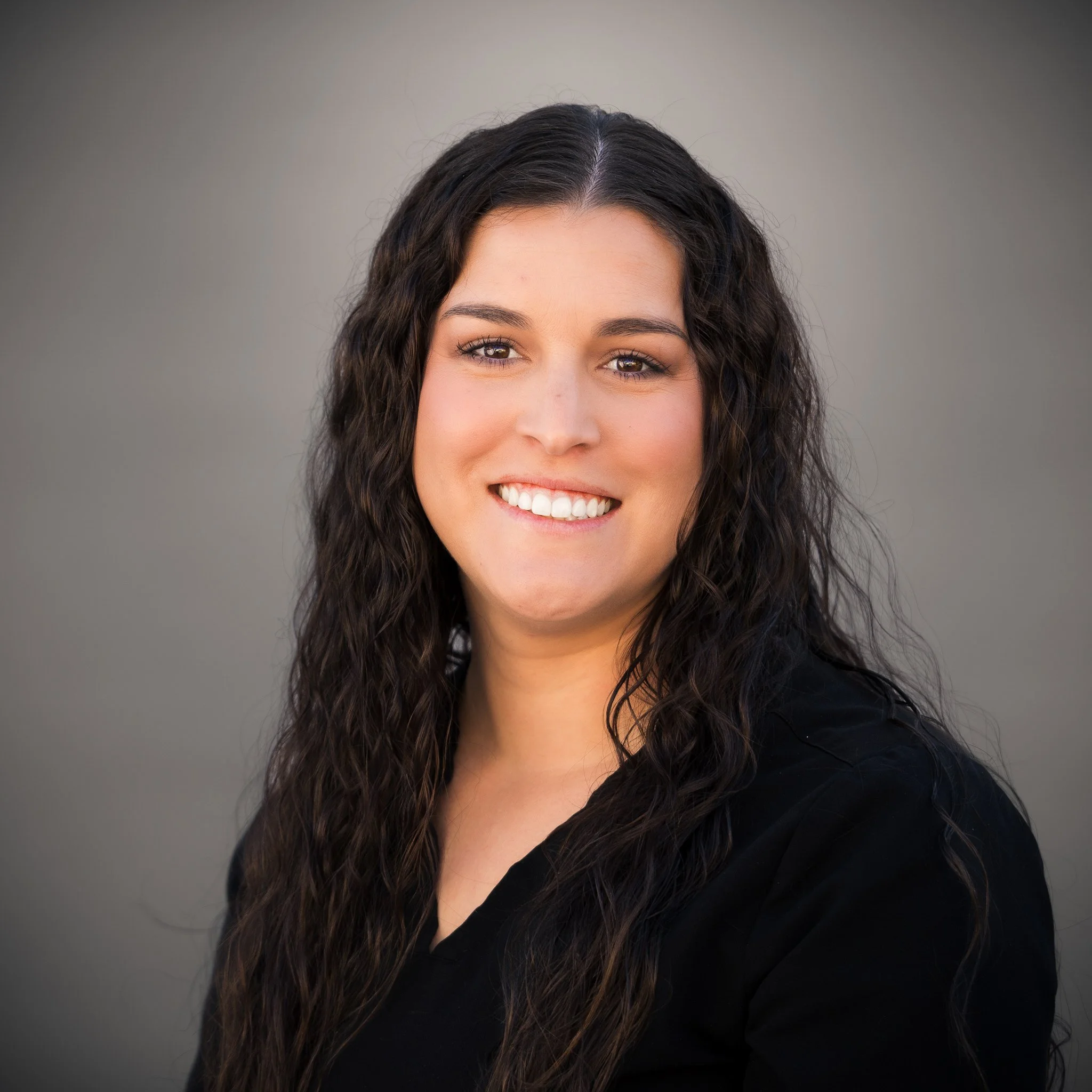 Smiling person with long brown hair wearing a black shirt against a plain background.