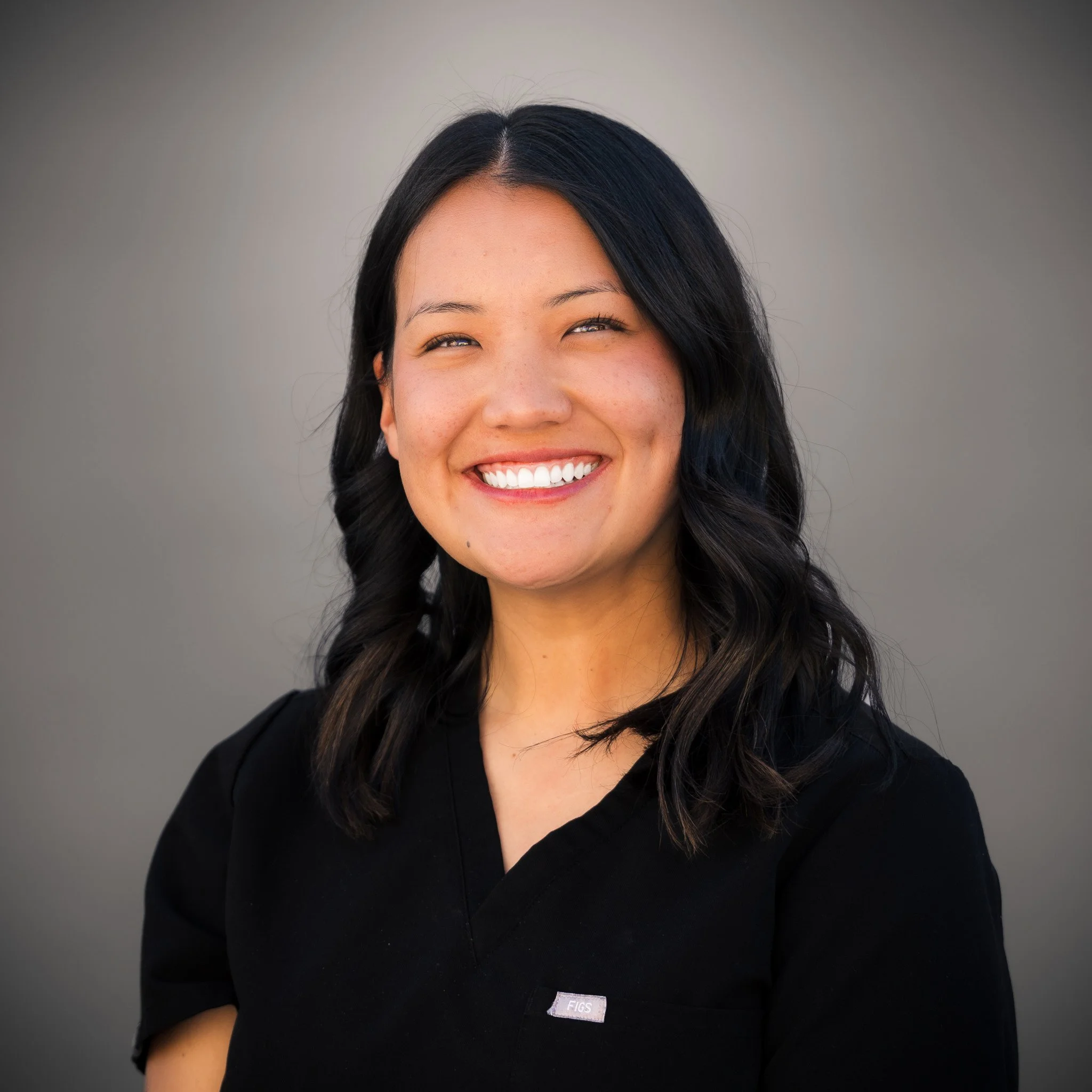 Young woman in black scrubs standing outdoors, smiling with a hand on her hip against a brick wall.