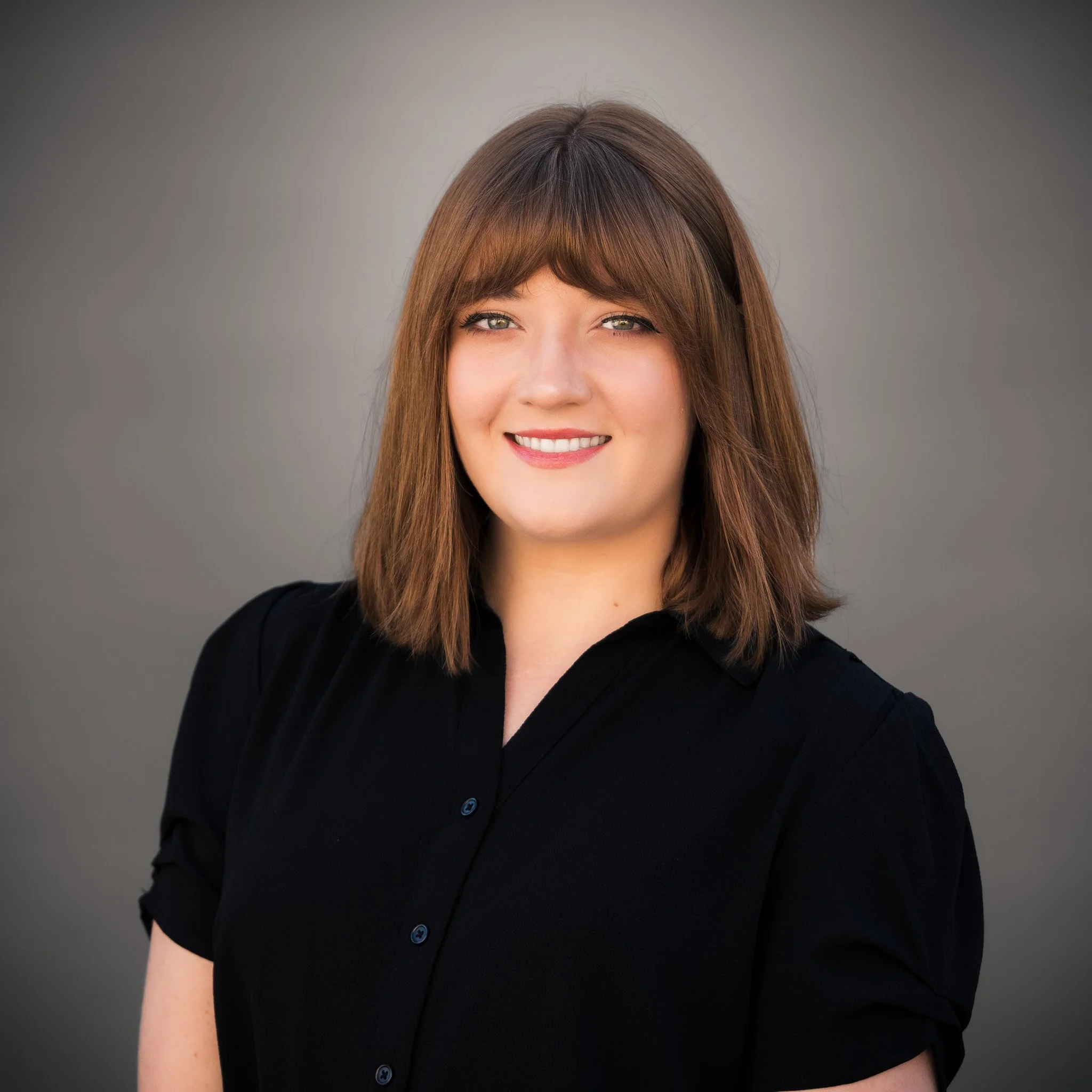 A woman with long red hair smiling outdoors with green trees in the background.