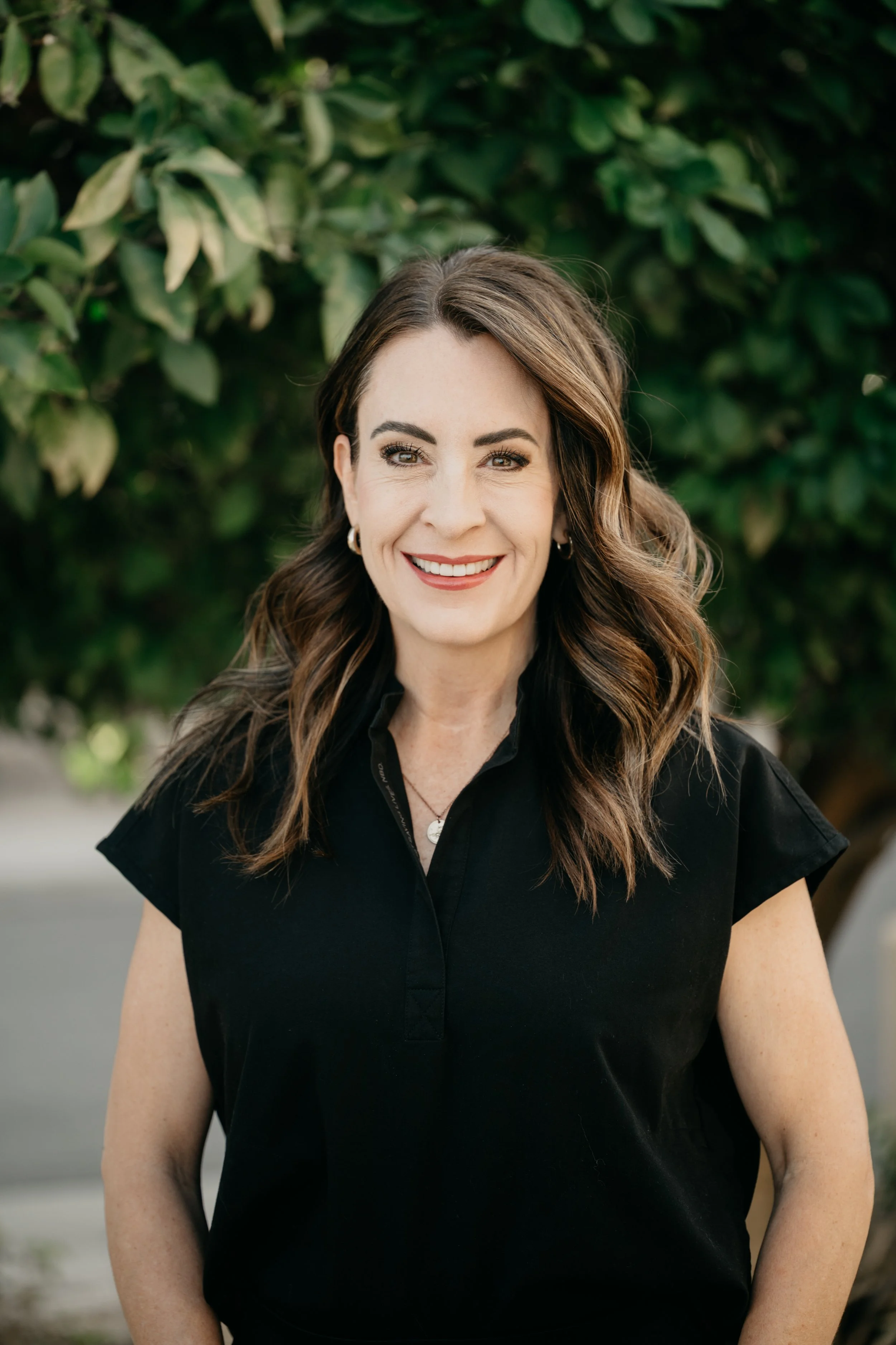 A woman with long brown hair, wearing a black dress, standing outdoors near a sidewalk with trees and a building in the background, smiling at the camera.