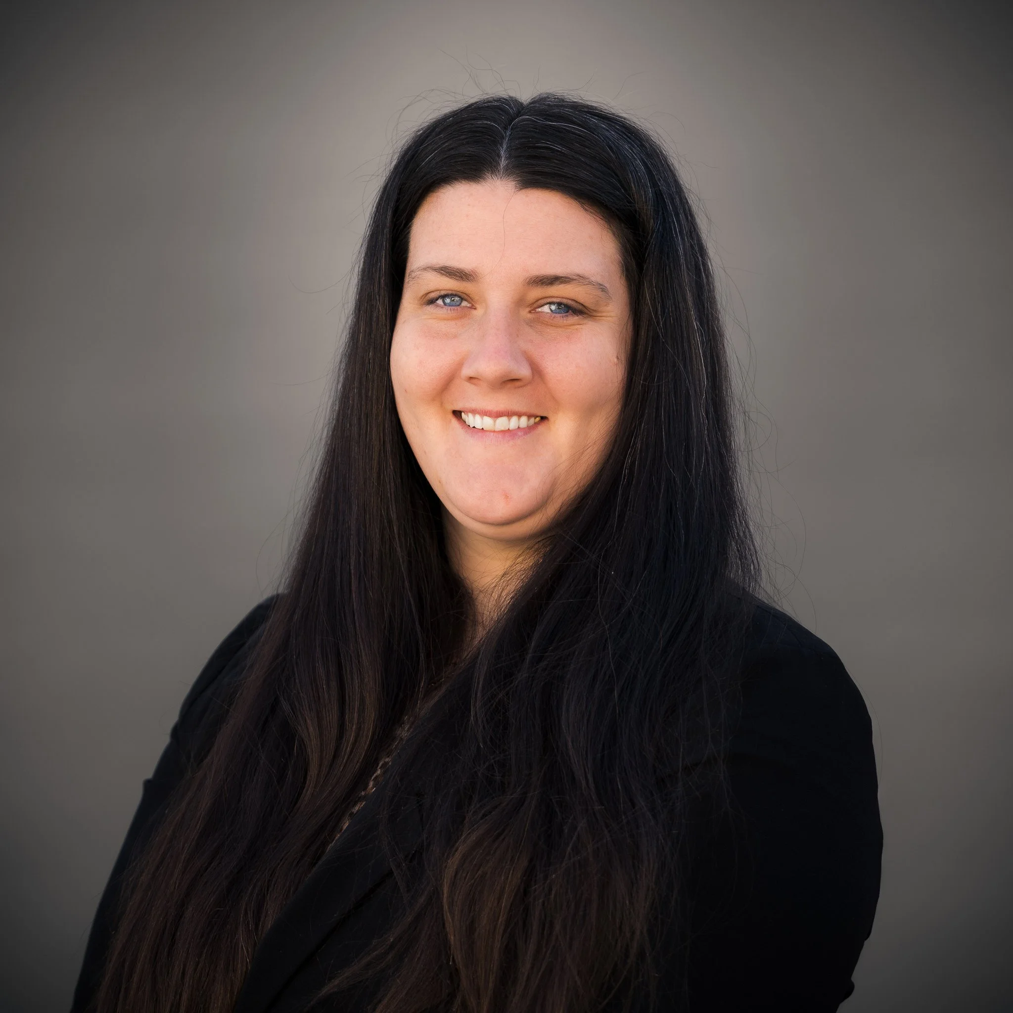 A smiling woman with long dark hair, wearing a black dress with sheer, dotted sleeves, stands outdoors in front of green trees and a partially visible brick building.