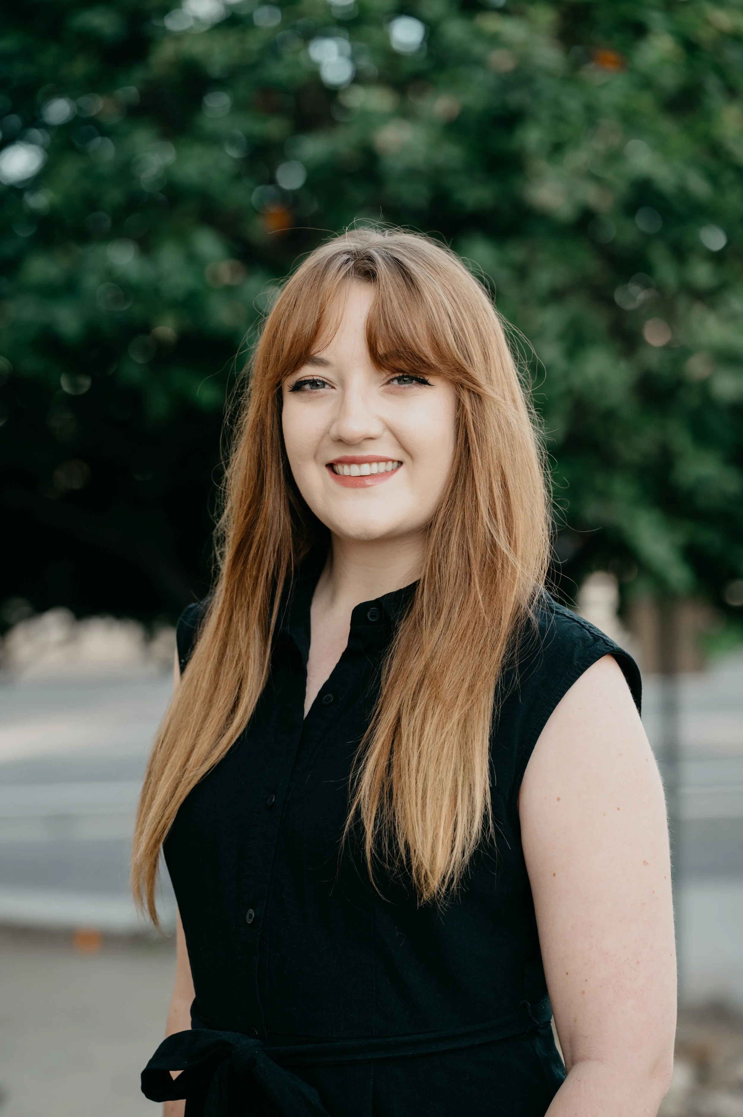 A woman with long red hair smiling outdoors with green trees in the background.