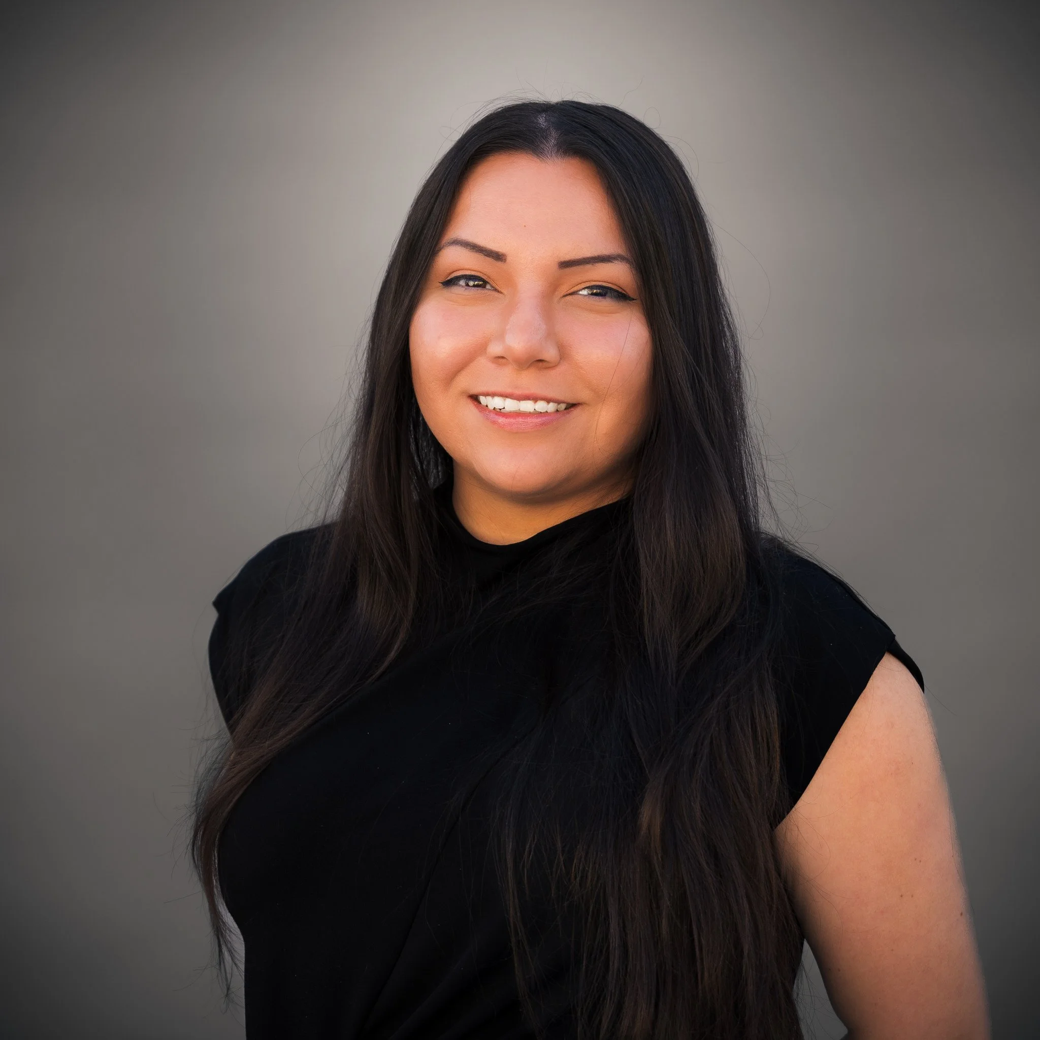 A woman with long dark hair, wearing a black t-shirt and gold necklace, standing outdoors with trees in the background.