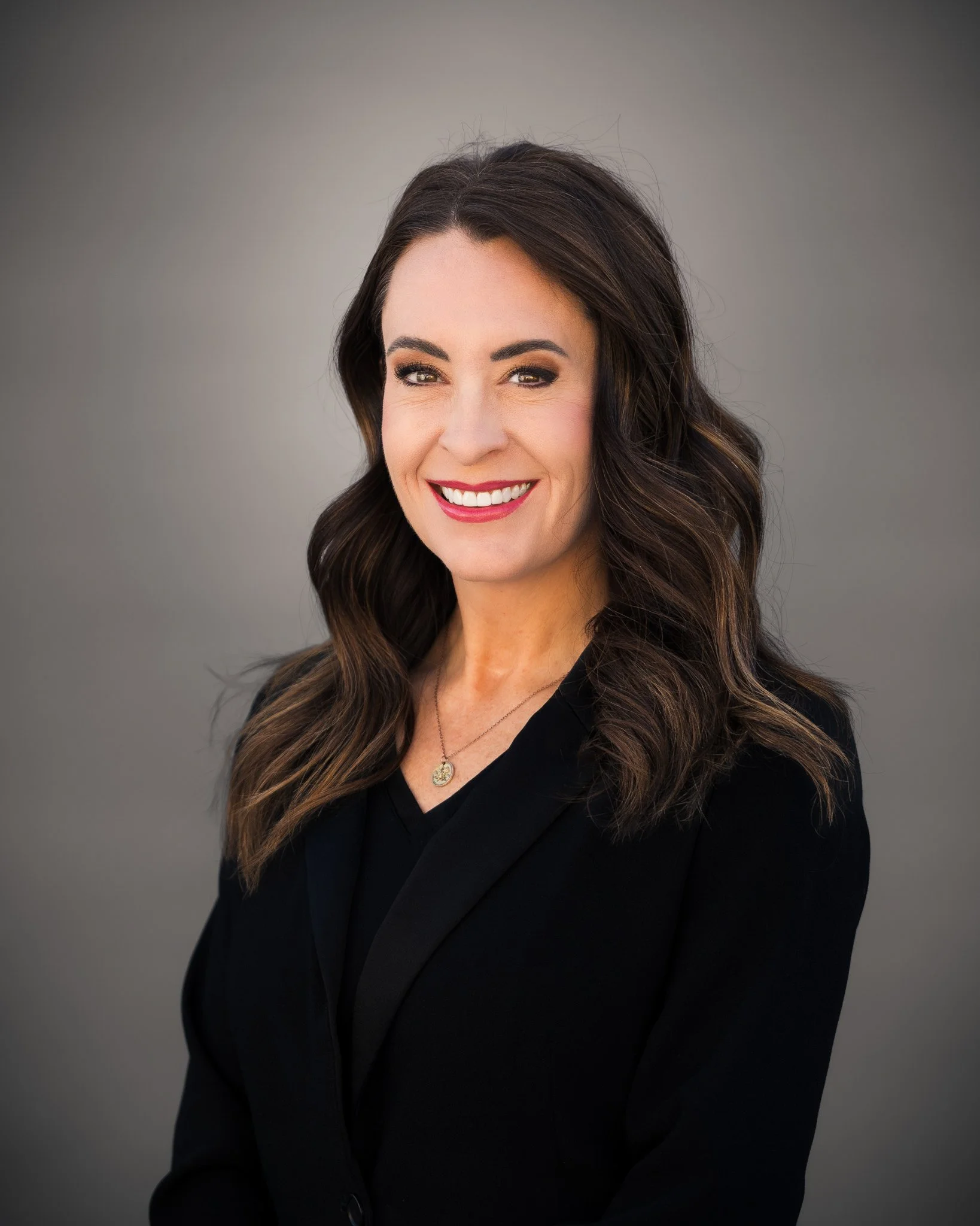 A woman with long brown hair, wearing a black dress, standing outdoors near a sidewalk with trees and a building in the background, smiling at the camera.