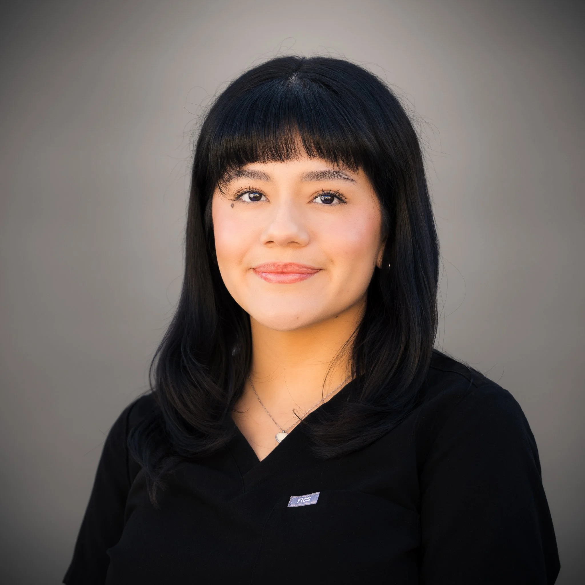 Smiling woman with long hair and a nose ring, wearing a black outfit, against a beige background.