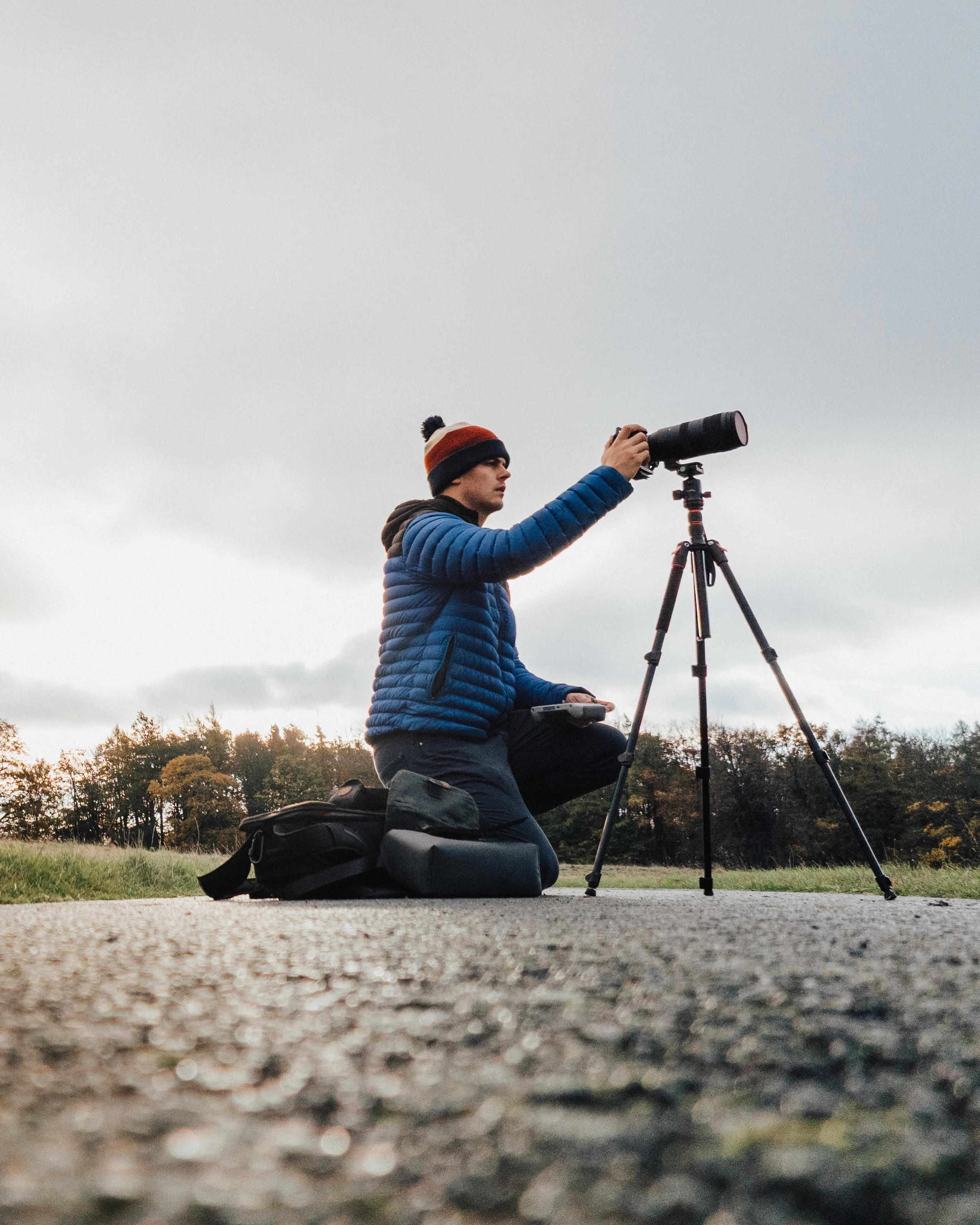 Person kneeling on ground, adjusting camera on tripod, wearing blue jacket and beanie, with camera bag nearby, outdoors.