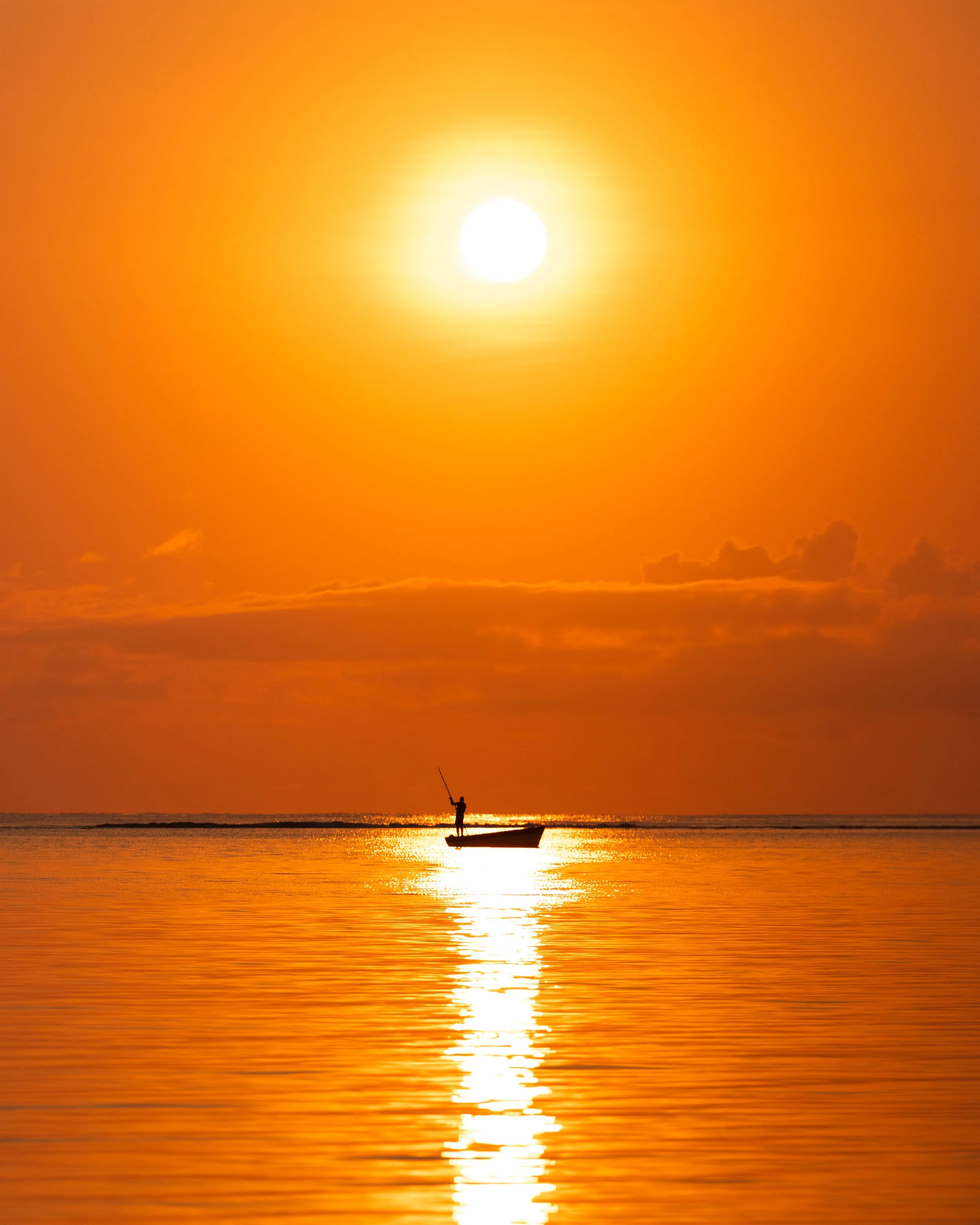 Silhouetted person in a small boat during orange sunset on calm water.