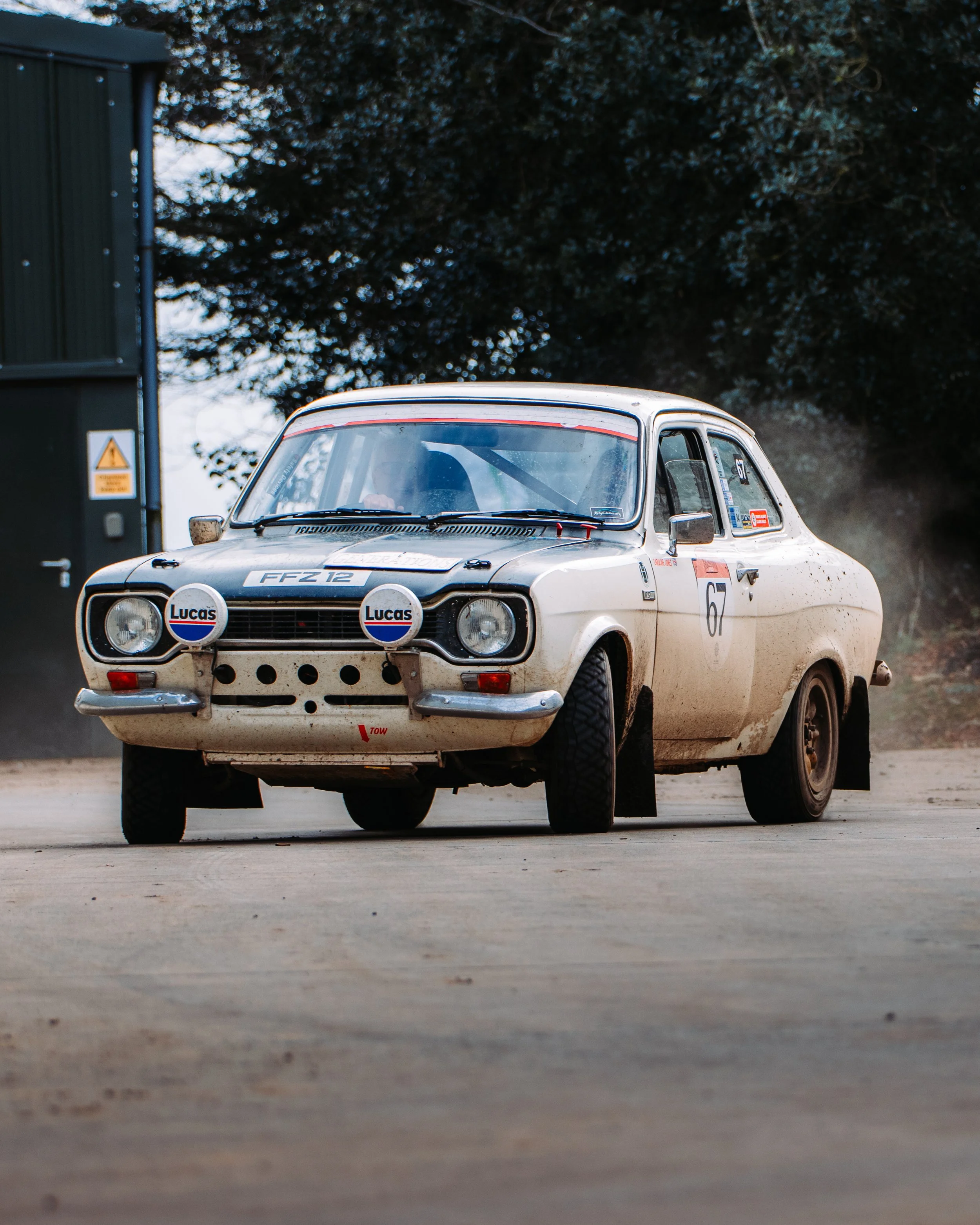 White vintage rally car with number 67 in motion on a dirt road, featuring front decals and Lucas lights, surrounded by trees and building.