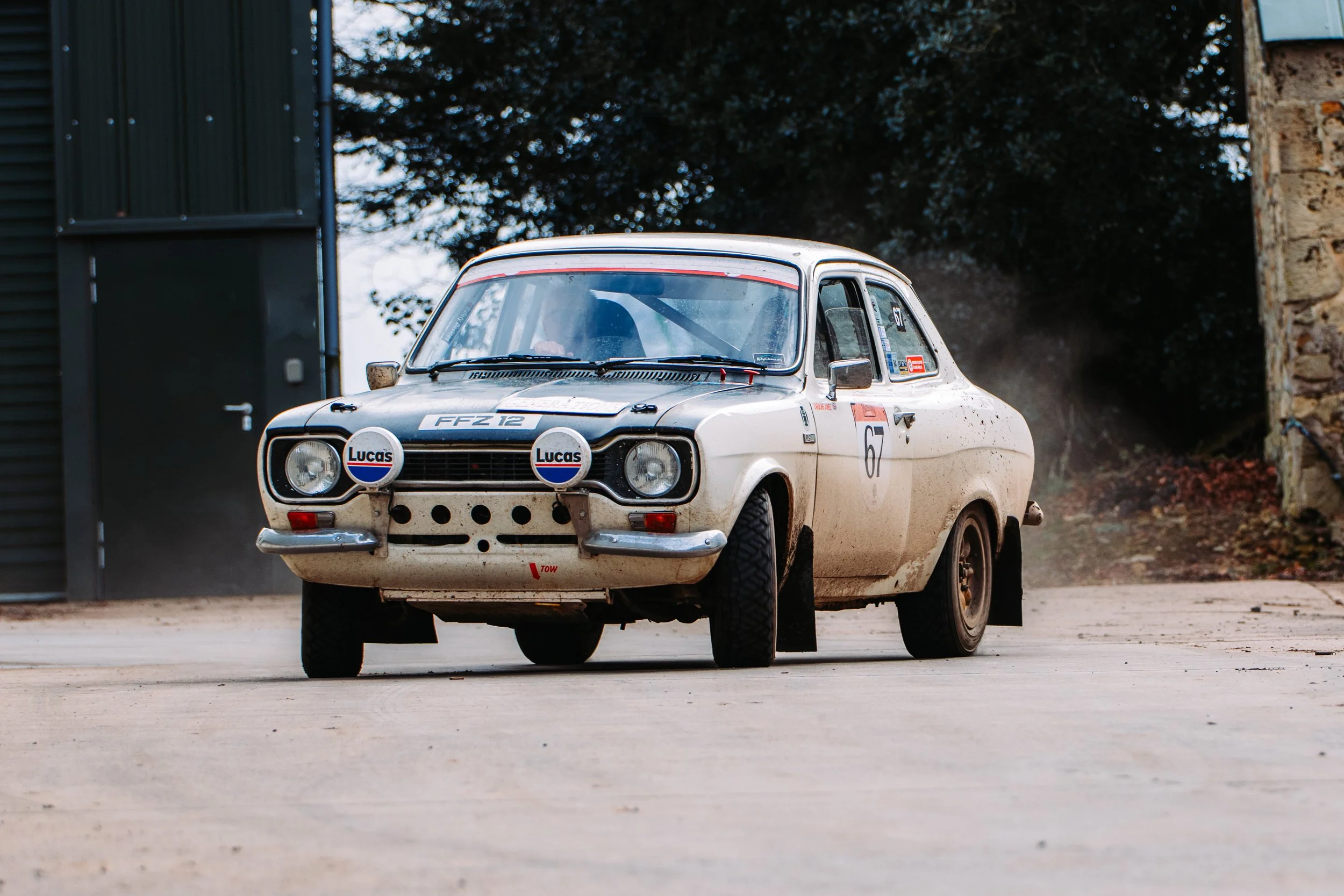 Vintage rally car with decals and mud on the body, parked near a building.