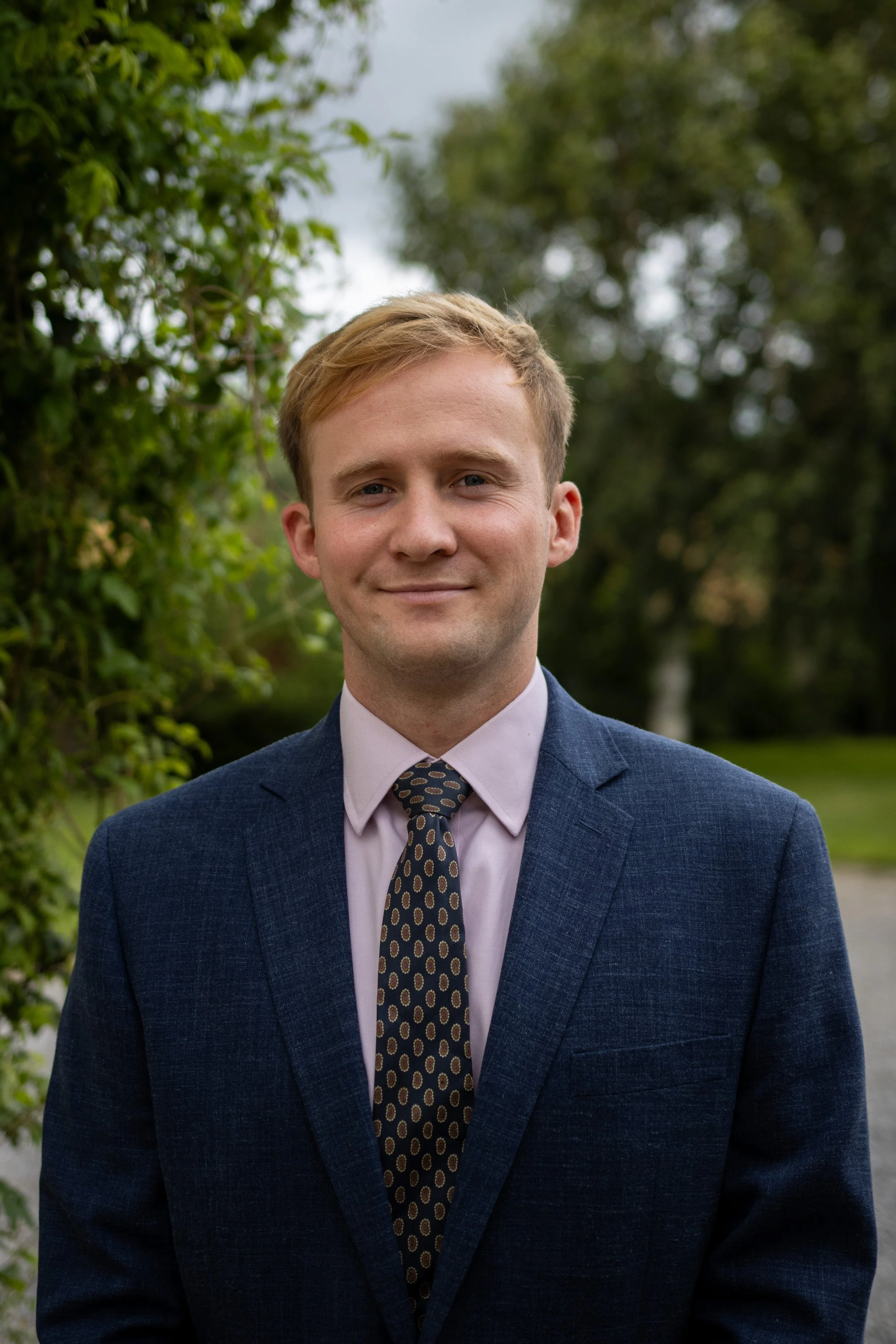 Man in a suit with a patterned tie standing outdoors in a garden setting.