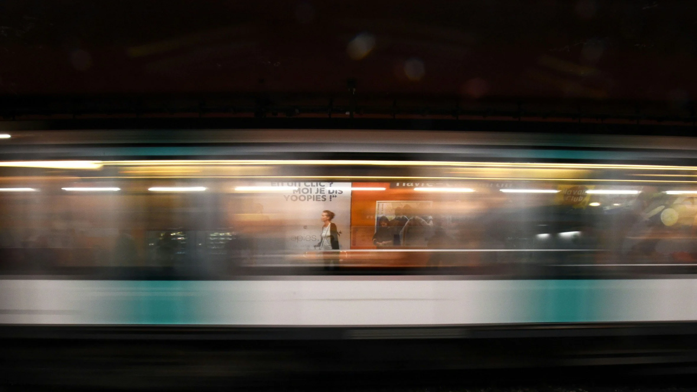 A train in motion at a station, with a blurred passenger walking past advertisements on the platform.