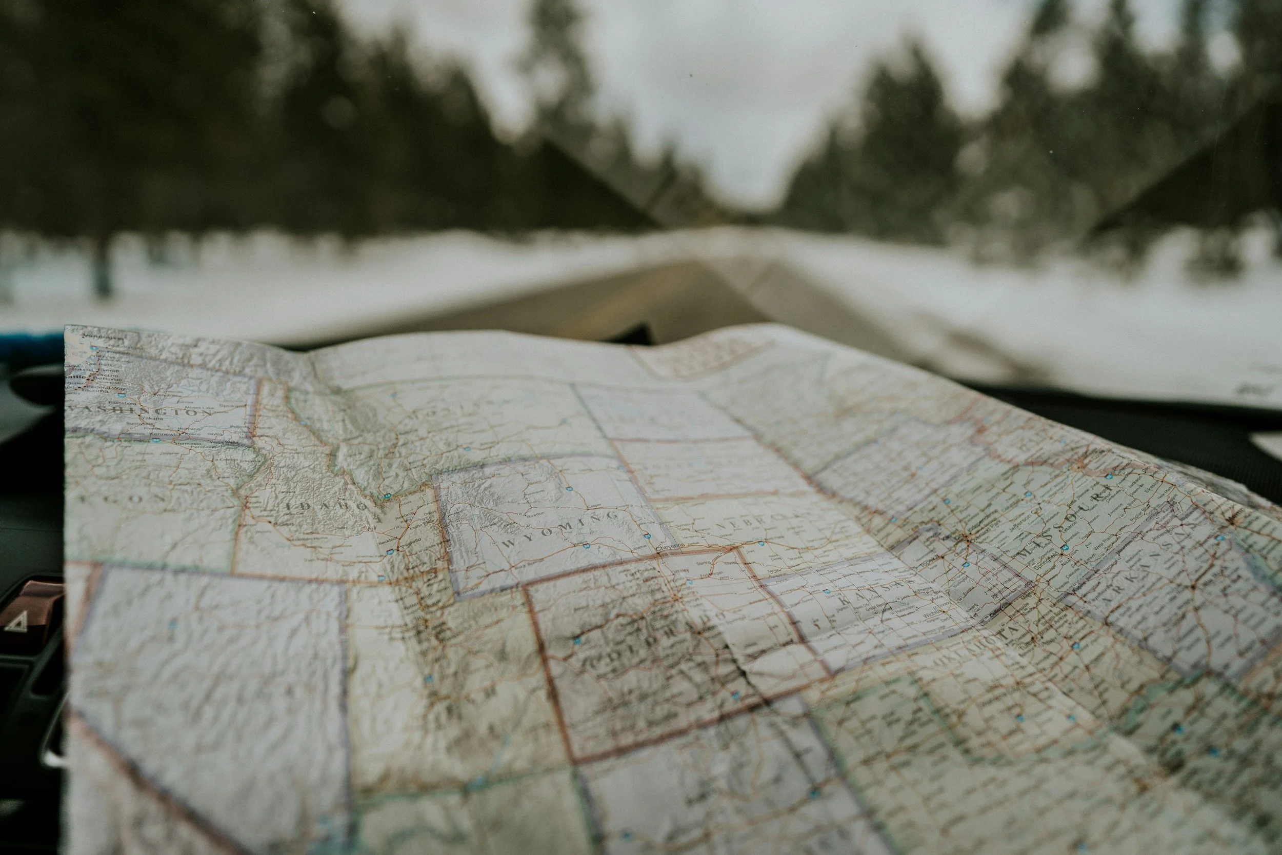A folded paper map of the United States on a car dashboard with a snowy landscape visible through the windshield.