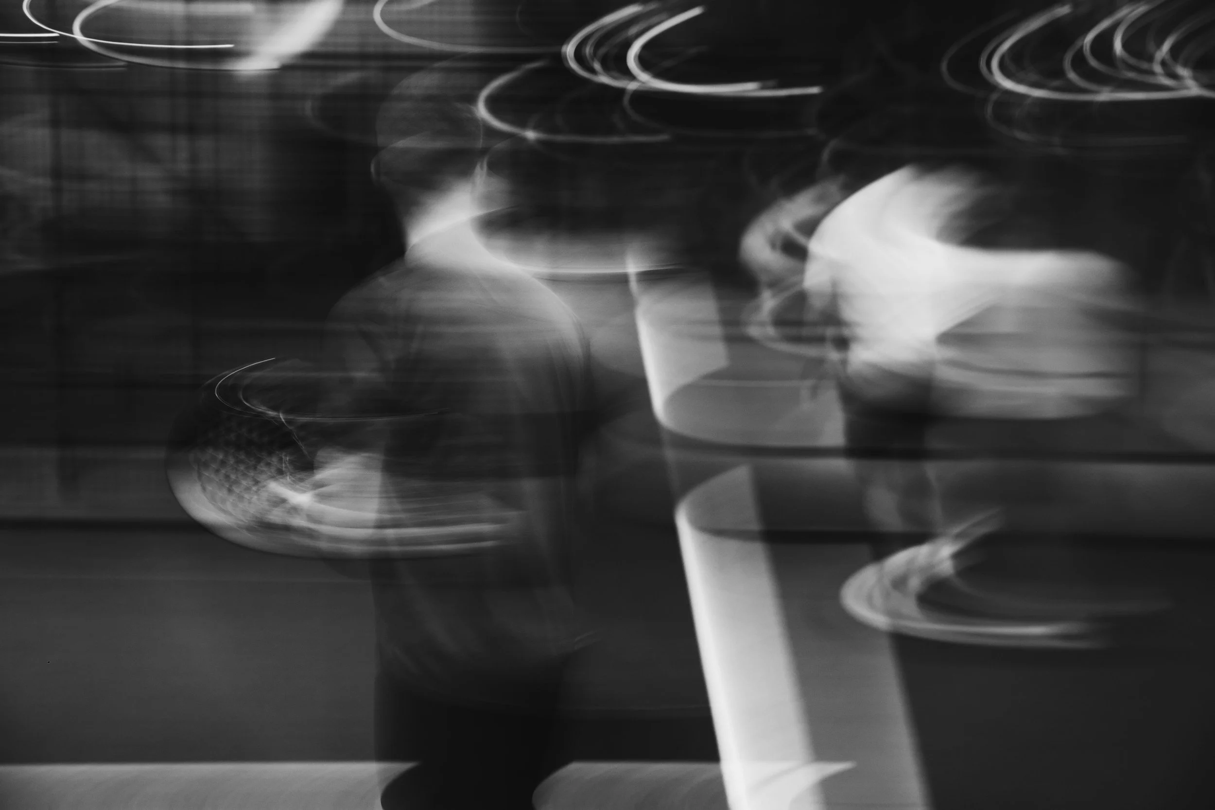 Blurred black and white photo of a person walking past a lighted area, with circular light streaks and motion blur.