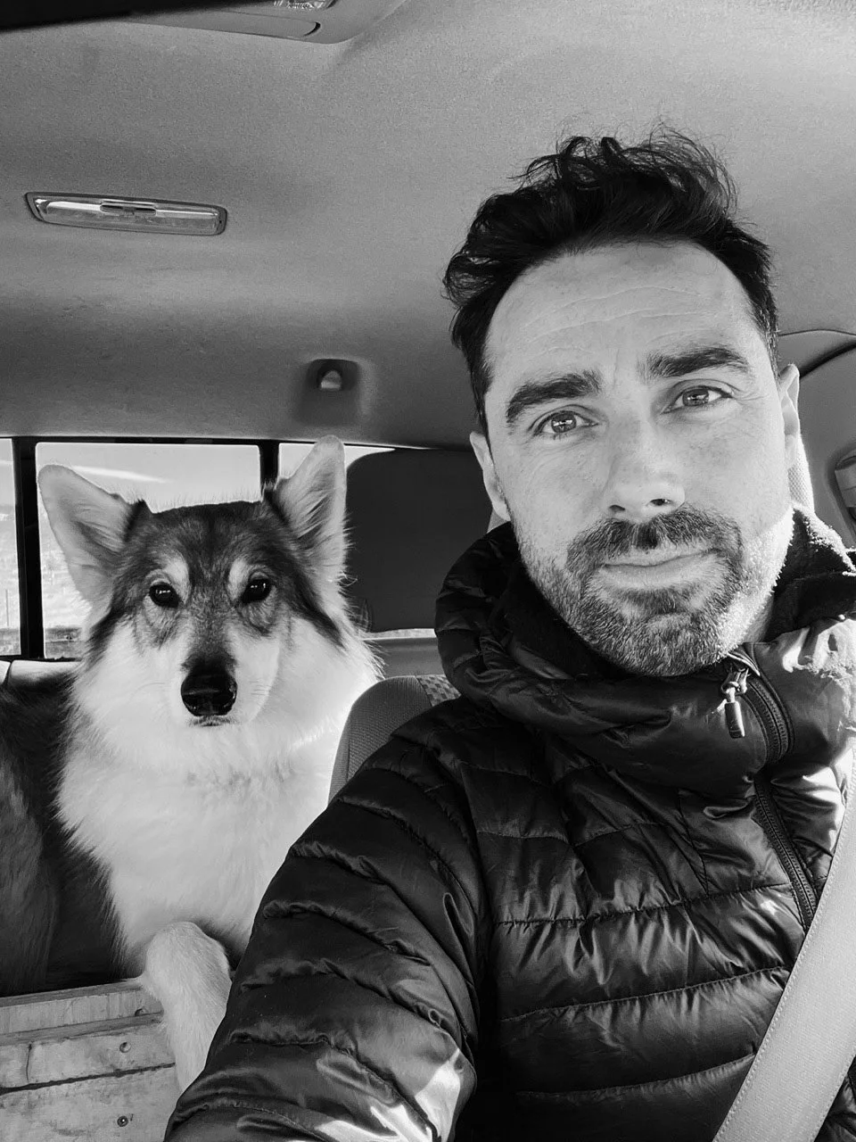 A man taking a selfie with his dog inside a car, both looking at the camera. The photo is in black and white.
