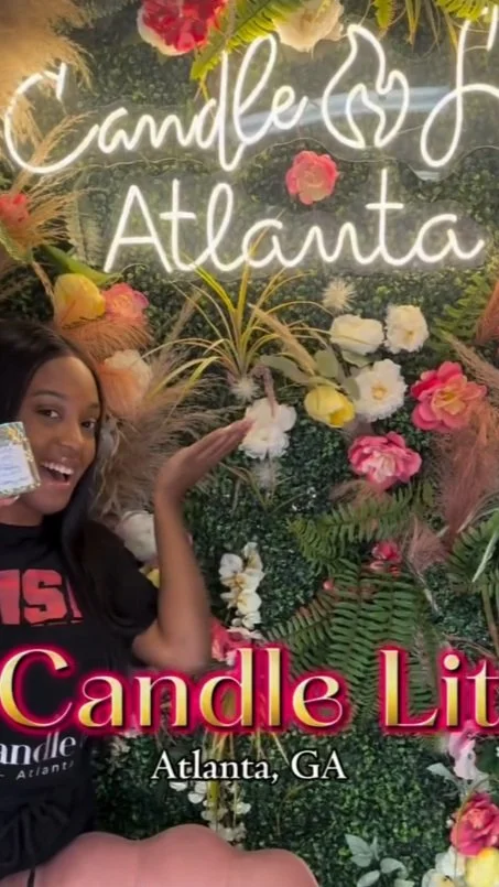 A woman smiling and gesturing towards a floral background with neon signs that read 'Candle Atlanta' and 'Candle Lit' in pink. The location is Atlanta, GA.