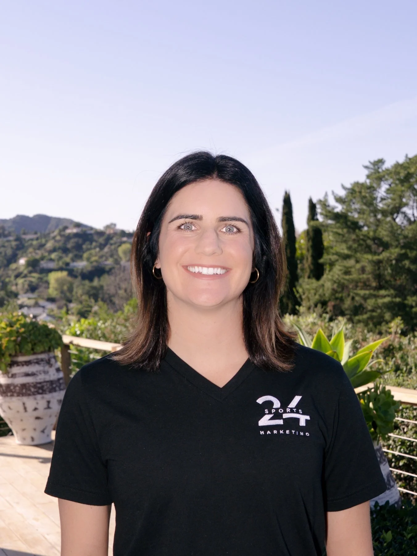 A woman with shoulder-length brown hair and blue eyes smiling, wearing a black shirt, with a blurred outdoor background.