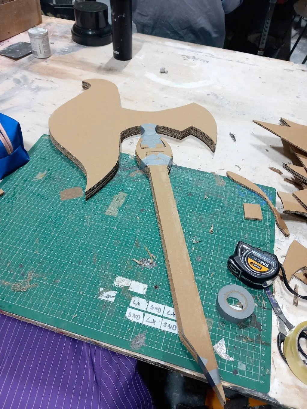 Workstation with a layered cardboard axe model, cutting mat, measuring tape, and tools.