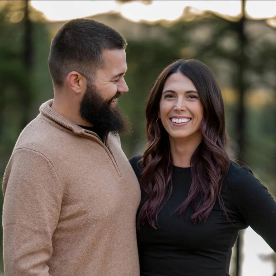 A smiling couple posing outdoors with trees in the blurred background. The man has a beard and wears a light brown sweater, while the woman has long dark hair and wears a black top.