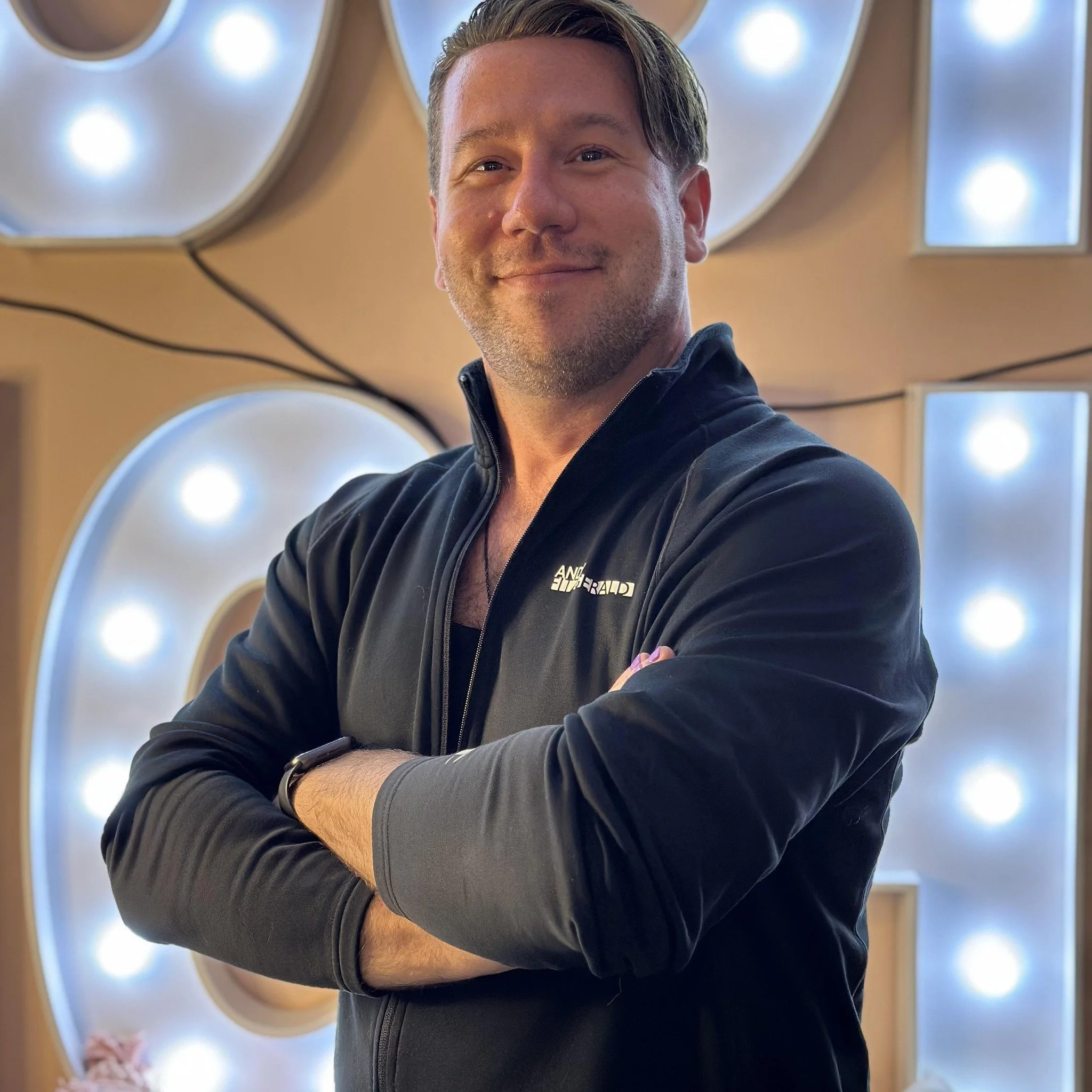 Person wearing a black jacket standing with arms crossed in front of illuminated background.