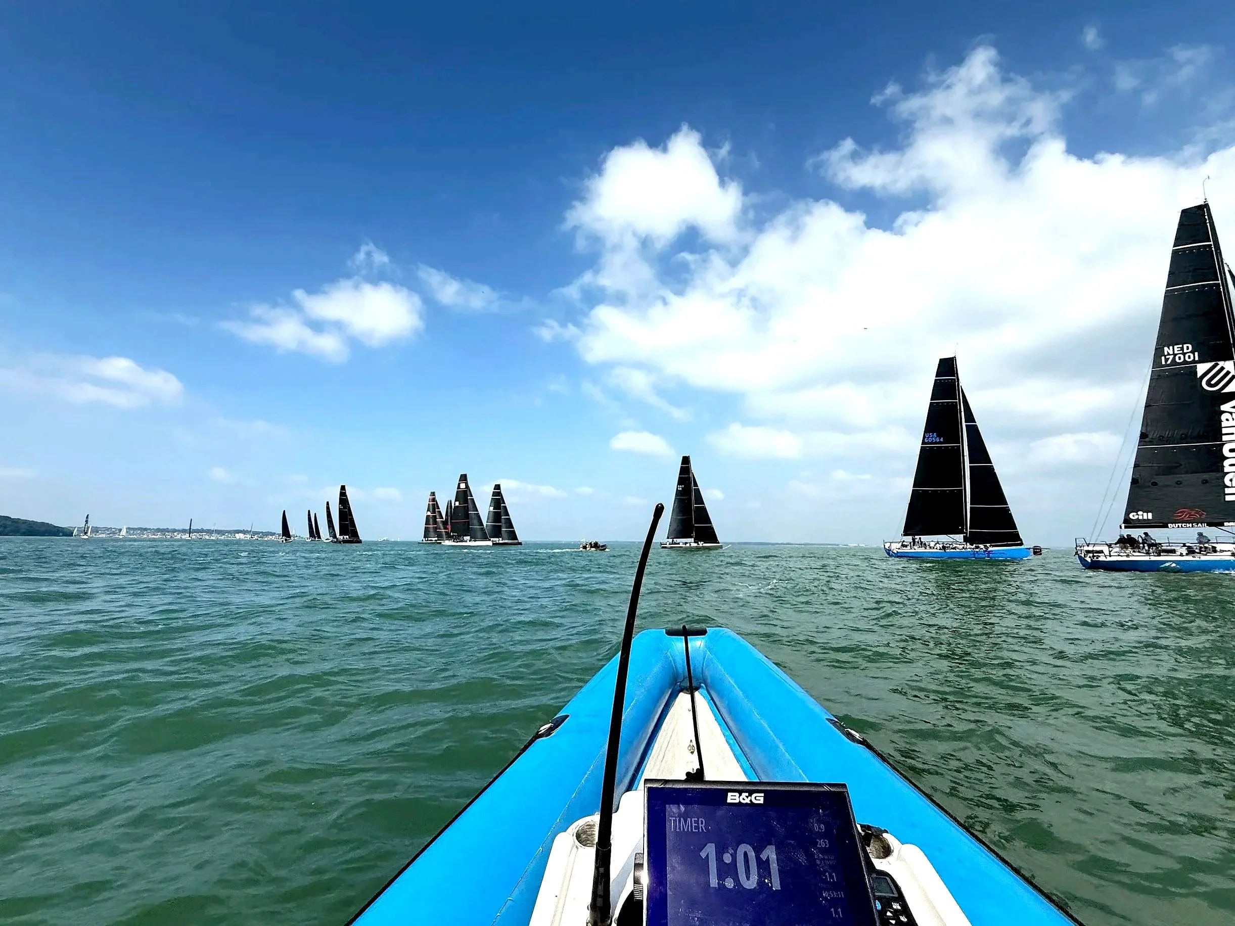 View from a boat with a blue and white bow on the water, showing a digital timer on a device reading 1:01, with several sailboats with black sails racing in the distance on a partly cloudy sky.