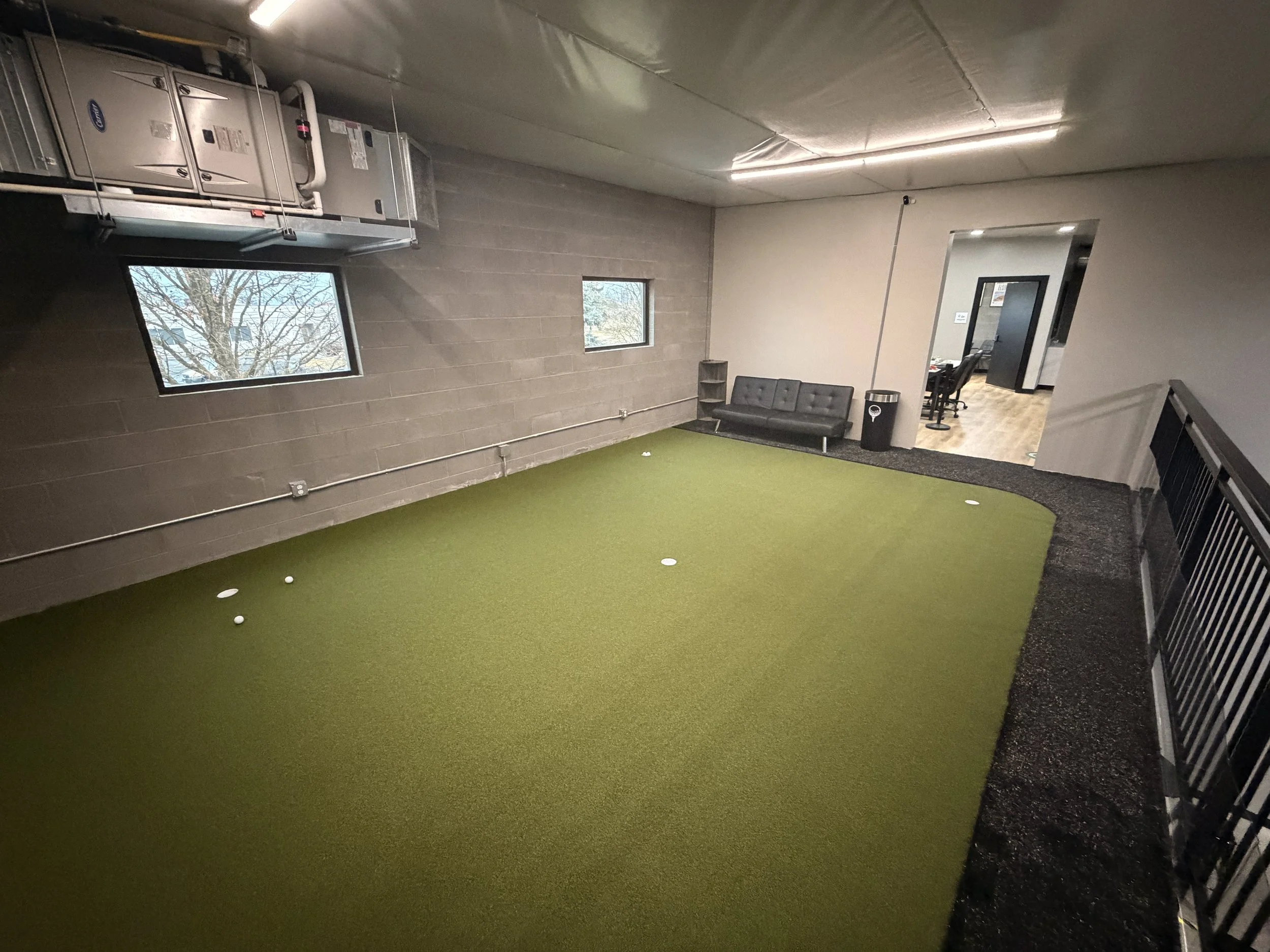Indoor golf practice area with artificial green turf, two small windows, a black couch, trash can, and a doorway leading to another room with chairs and tables.