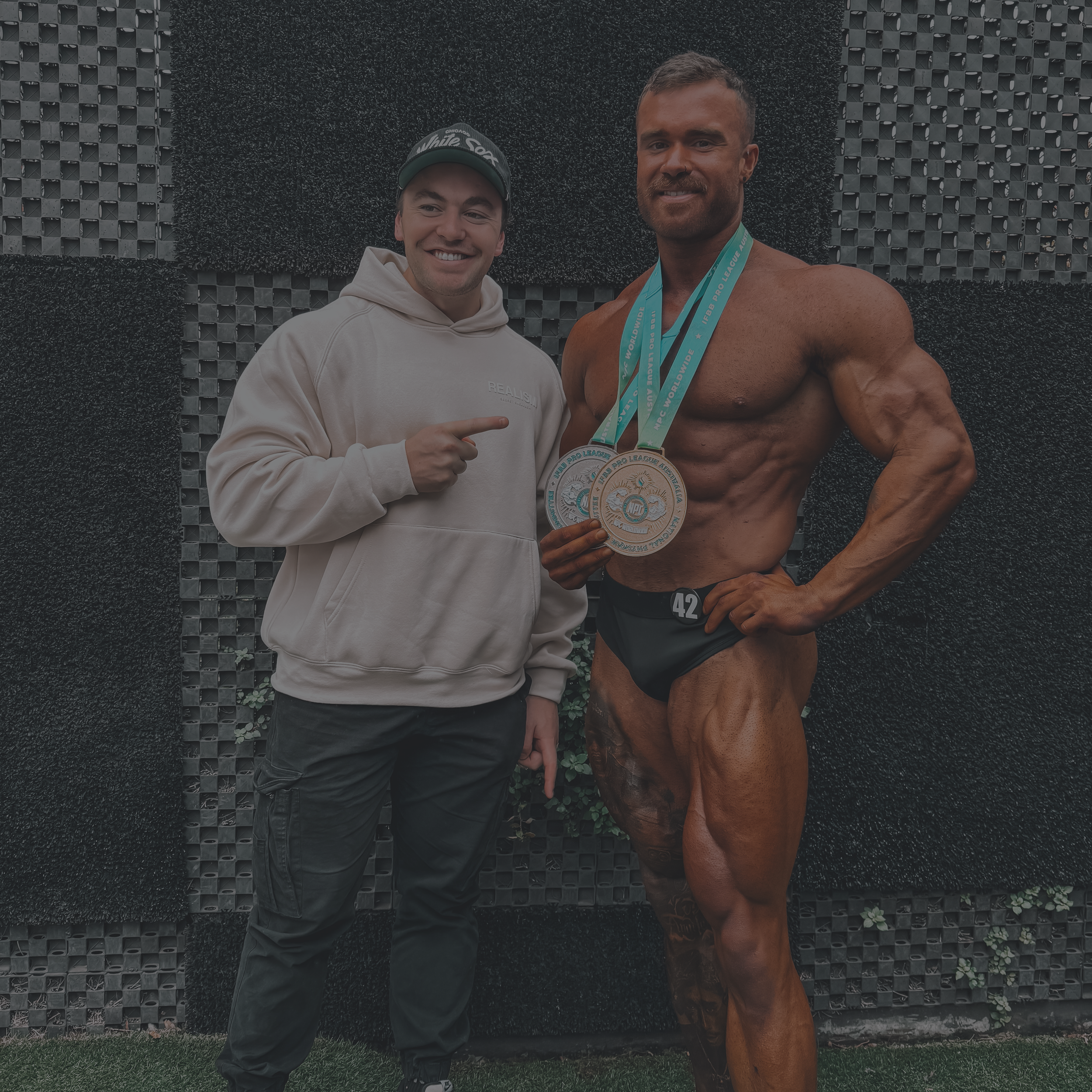 Sid and bodybuilder client with two medals, wearing casual attire, standing in front of a textured wall.