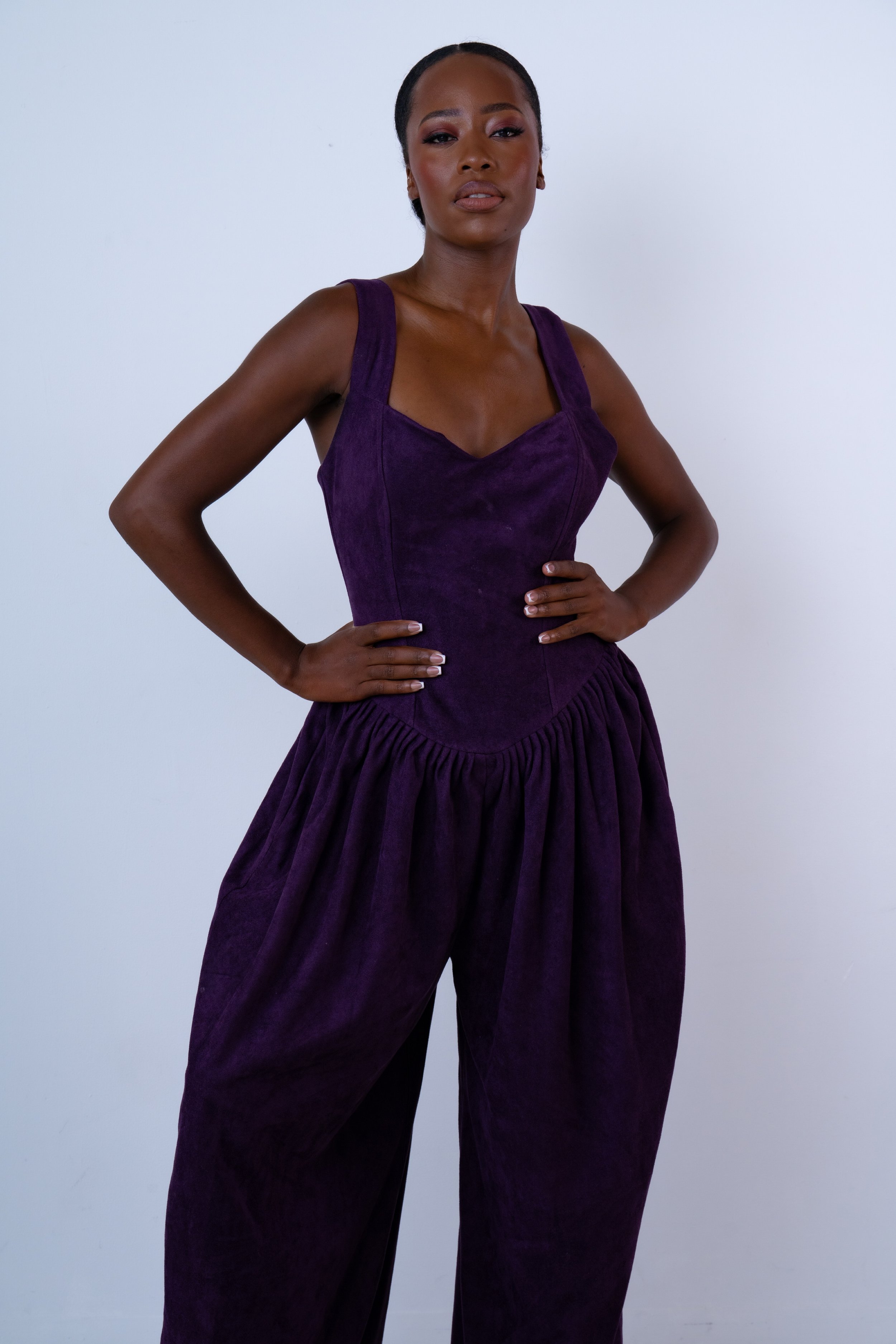 A woman wearing a purple sleeveless dress with pleated wide-legged pants, standing confidently against a plain white background.