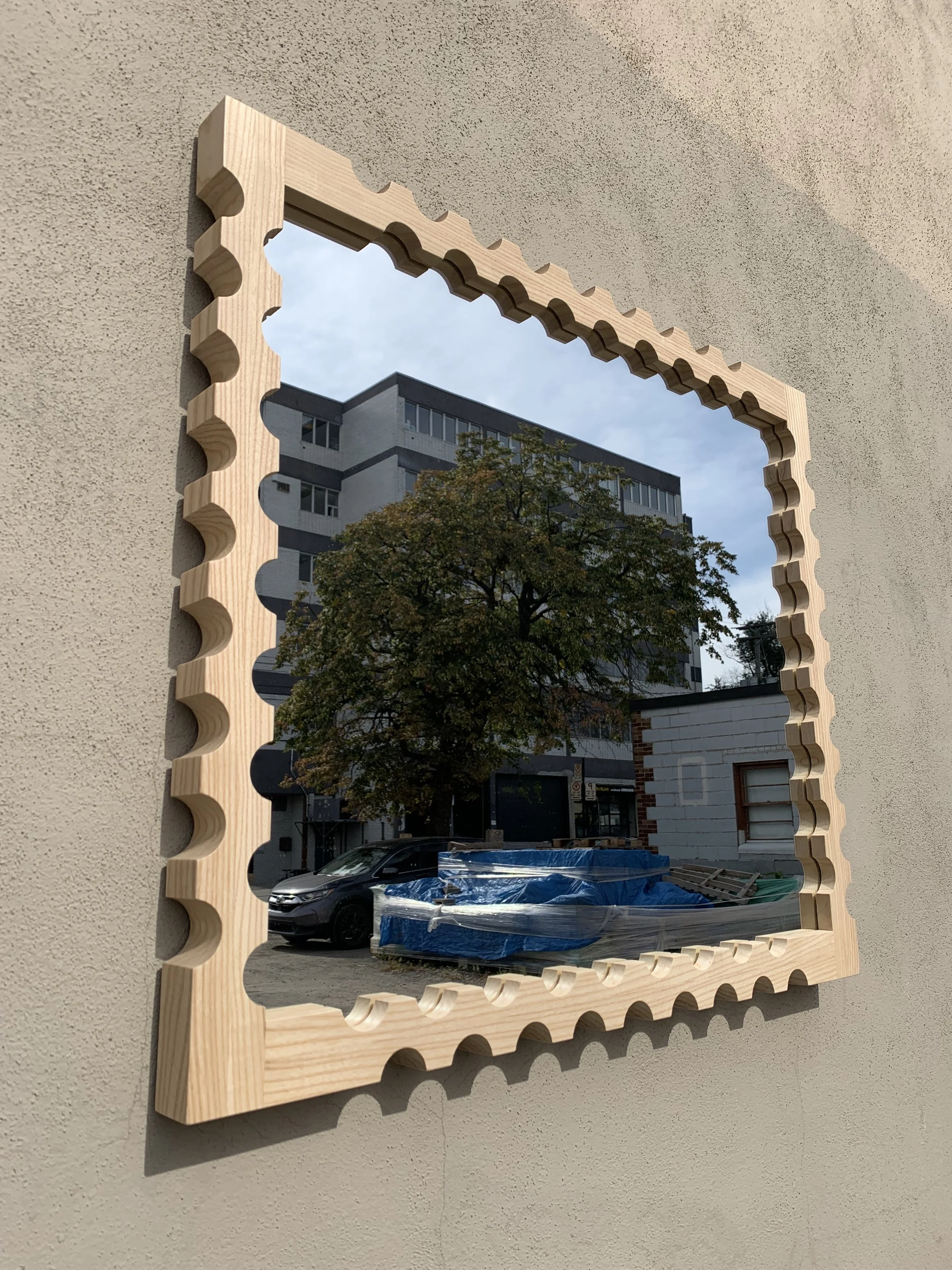 A rectangular mirror with a wooden frame featuring a scalloped edge design, mounted on a beige exterior wall. The mirror reflects an urban scene including a tree, a parked black car, a building, and a stack of blue items covered with plastic.