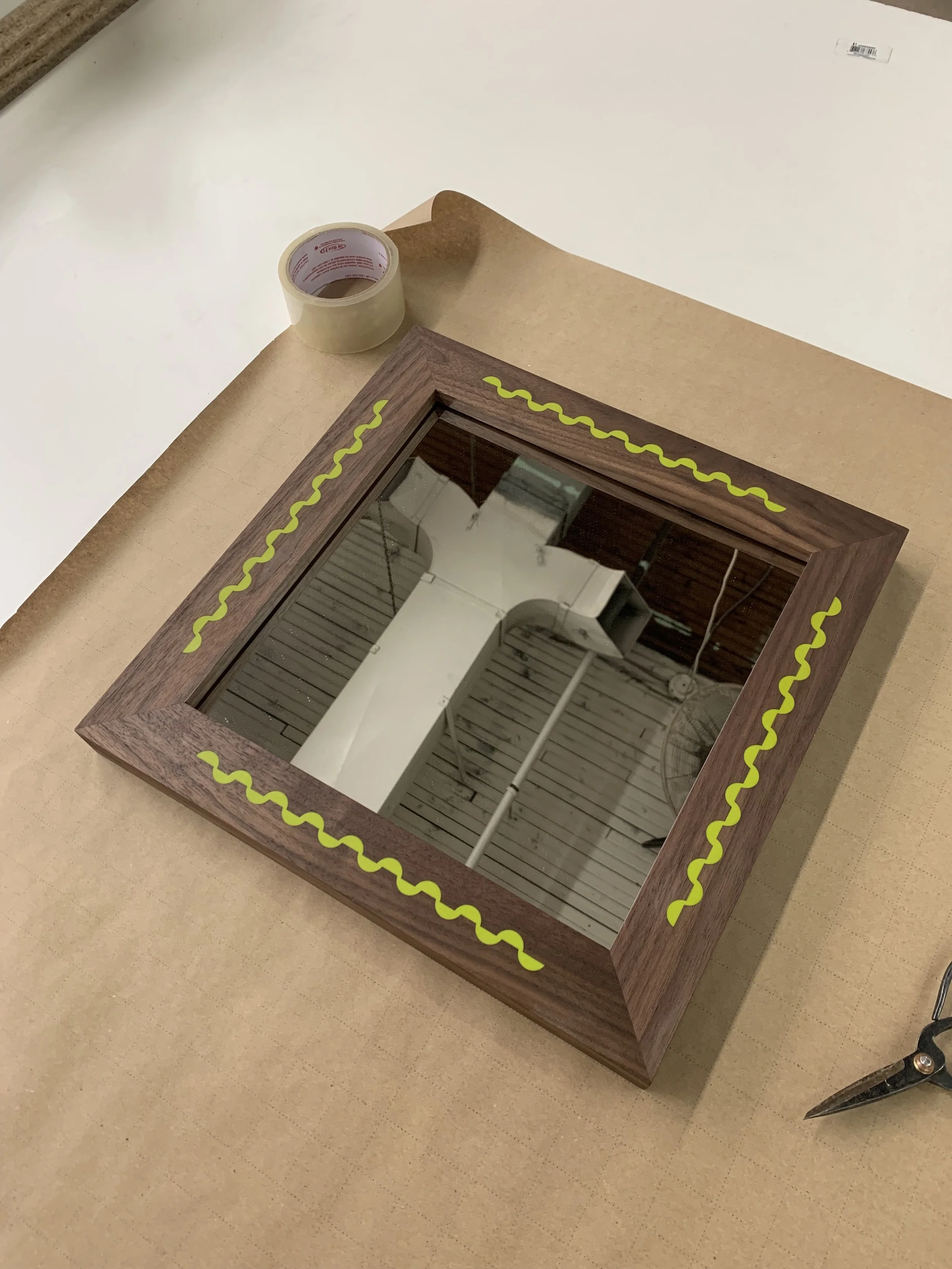 A mirror with a wooden frame decorated with yellow wavy lines, placed on a roll of brown craft paper, with a pair of scissors and tape nearby.