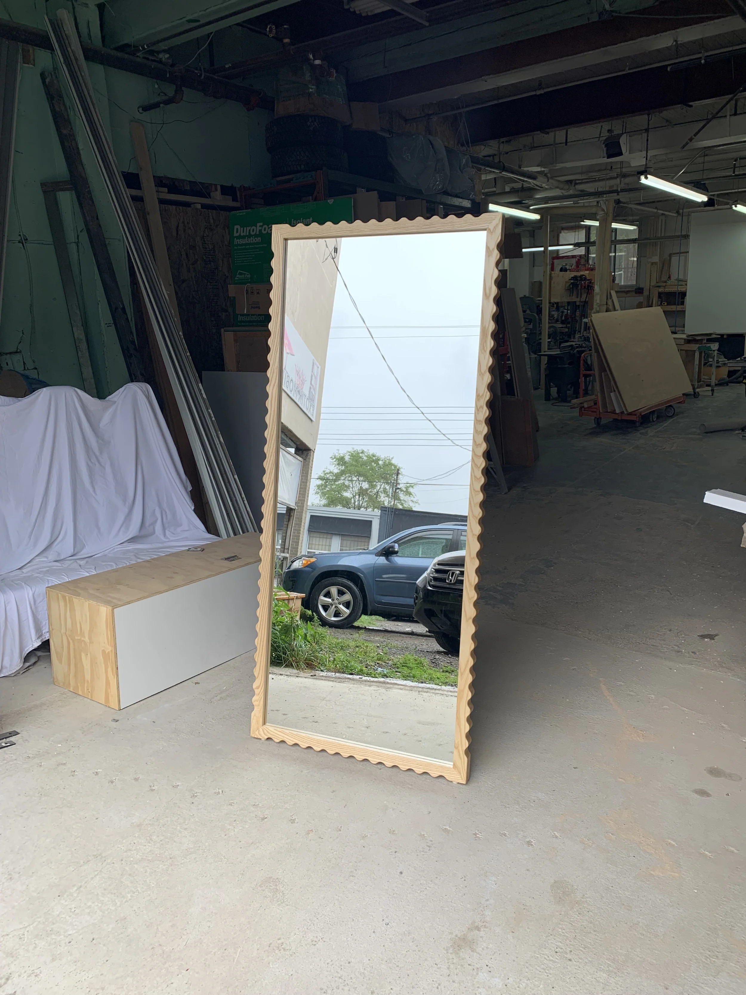 Full-length mirror with a scalloped wooden frame leaning against the wall inside a woodwork shop. Outside view with parked cars and cloudy sky reflected in the mirror.