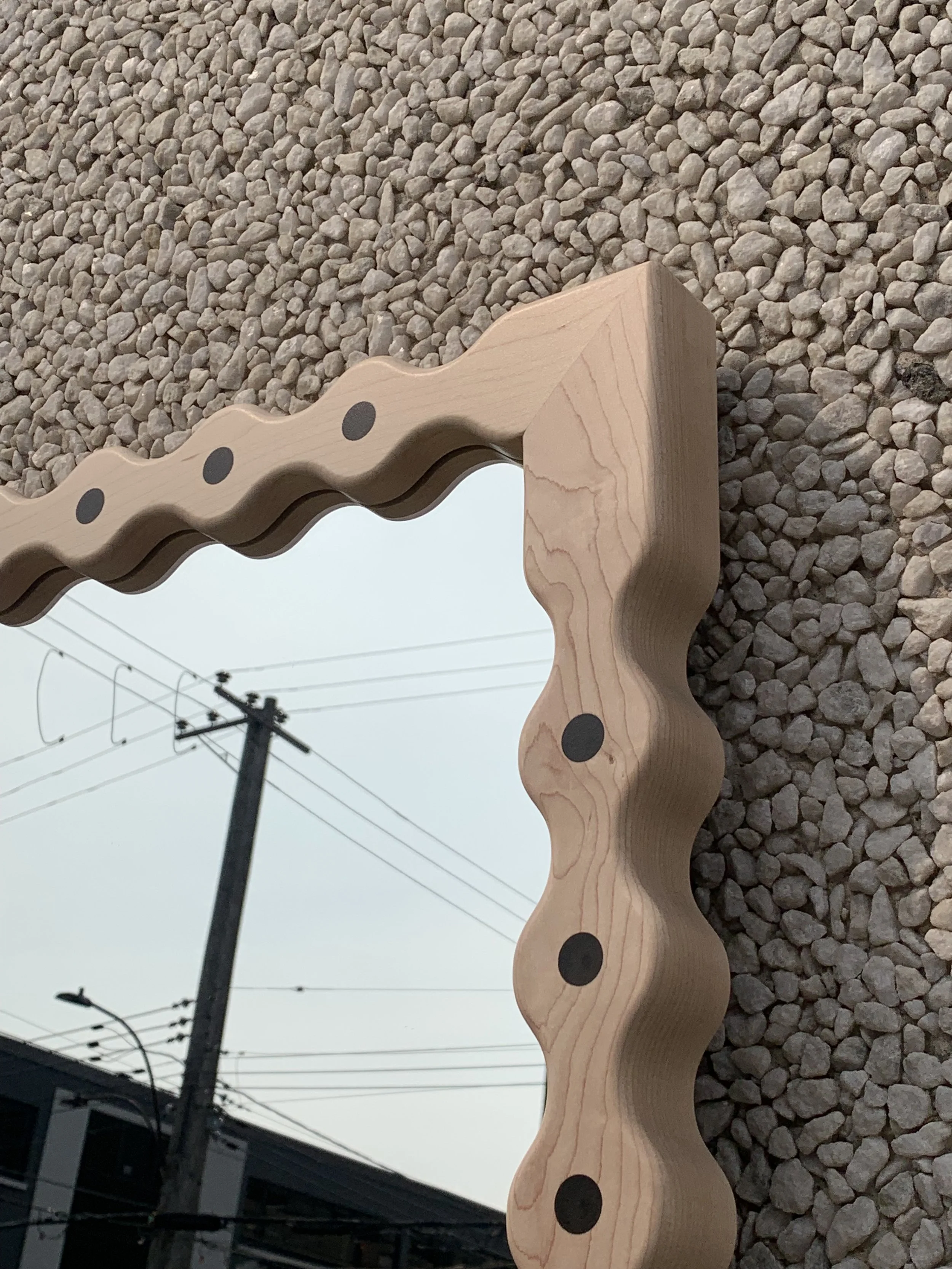 Close-up of a decorative mirror with a wavy wooden frame, placed on gravel with power lines and a pole reflected in the mirror.