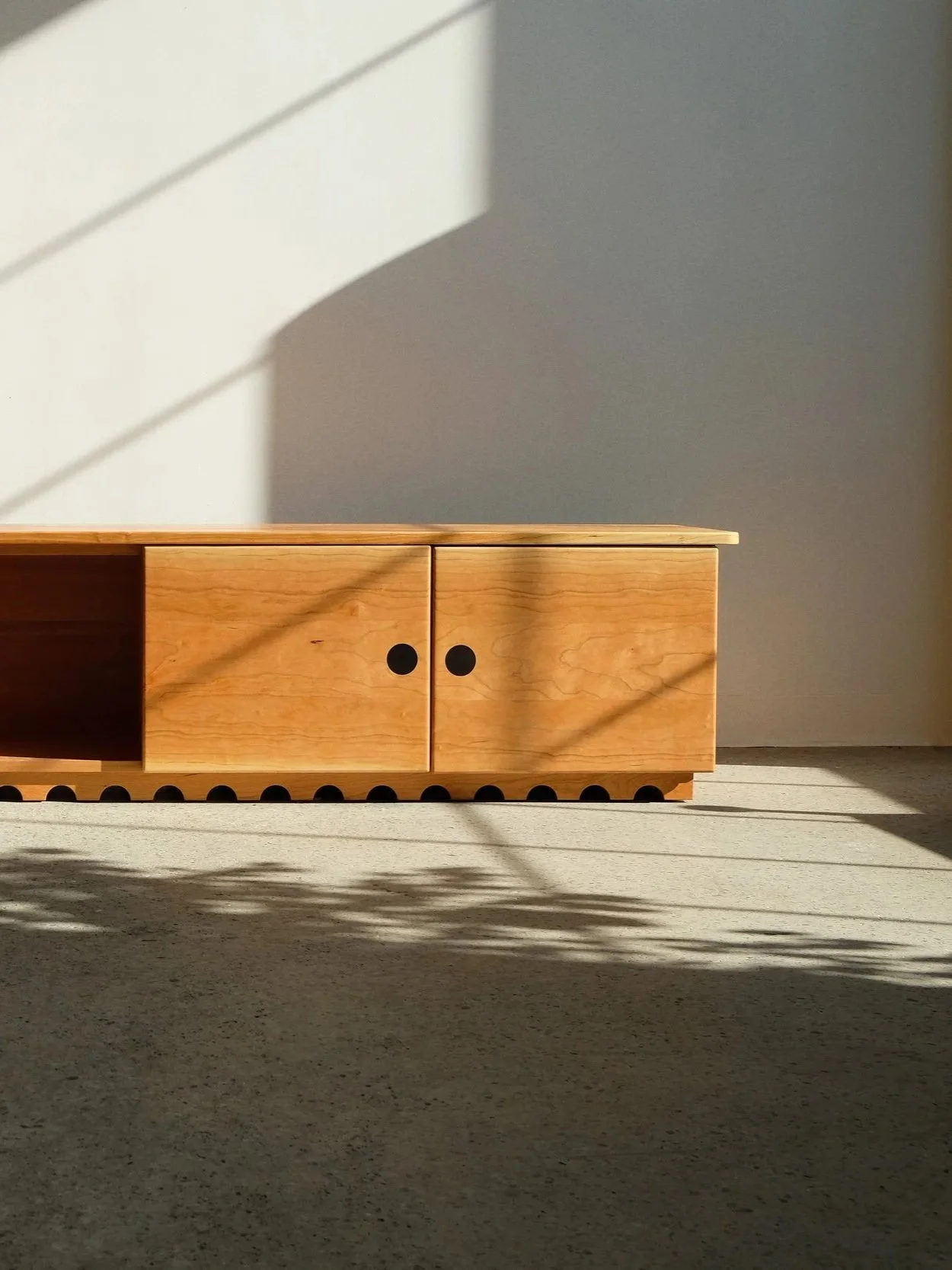 A wooden TV stand with black circular accents, sitting on a light-colored carpet with sunlight and shadows cast on it.