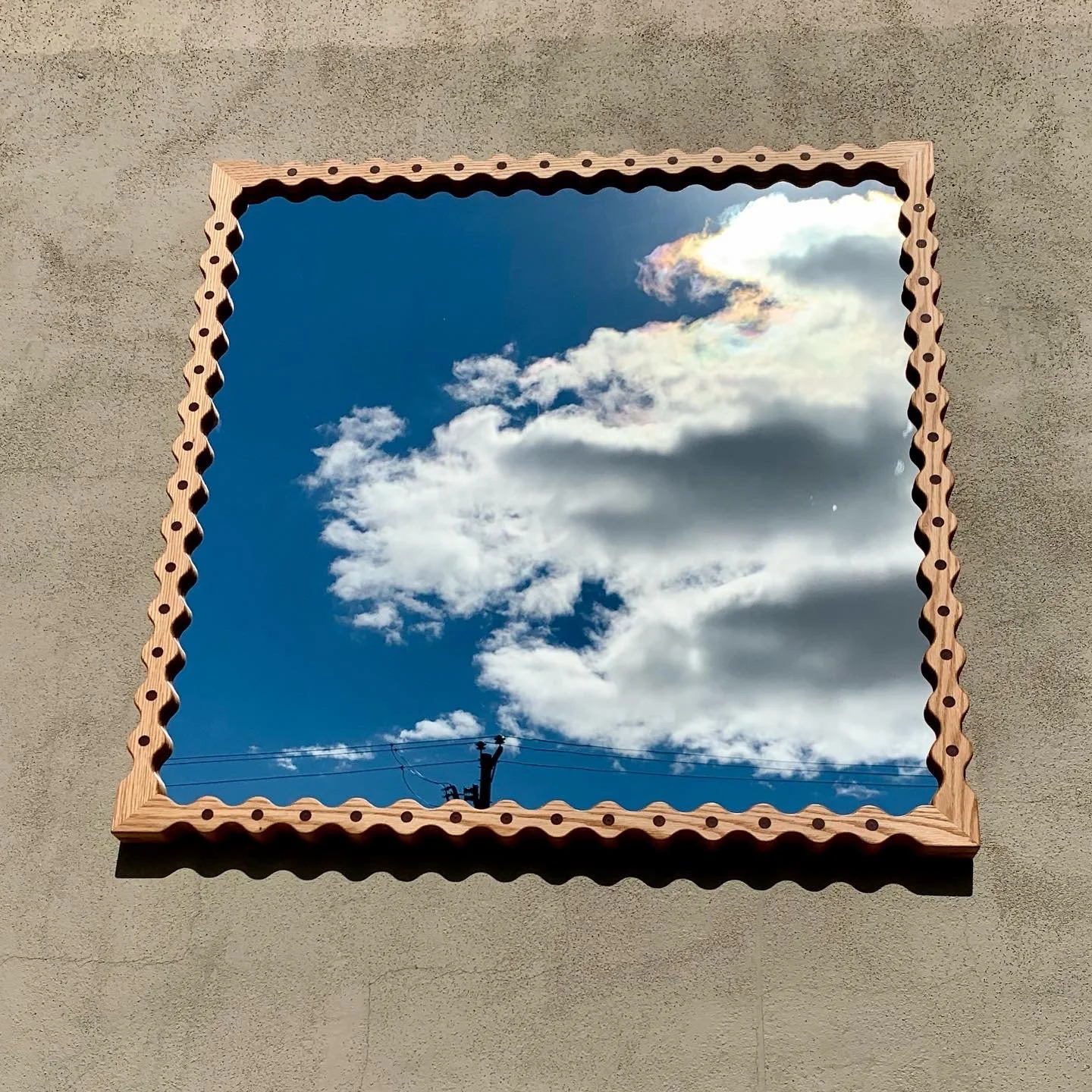 A rectangular mirror with a decorative wooden frame reflecting a blue sky with scattered white clouds and a utility pole with wires.