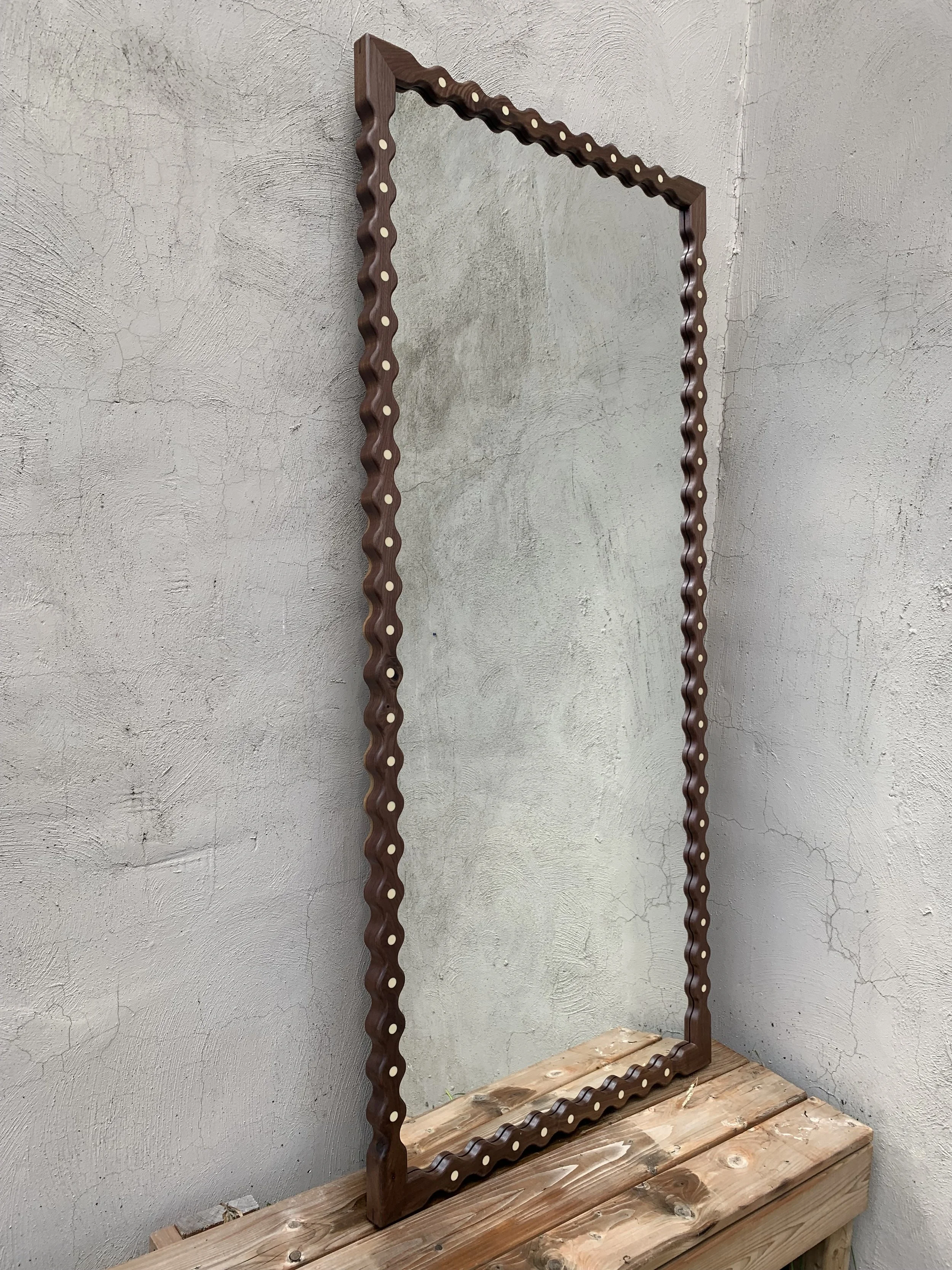 A wooden-framed mirror with a wavy, scalloped design along the edges, leaning against a textured gray wall on a wooden surface.