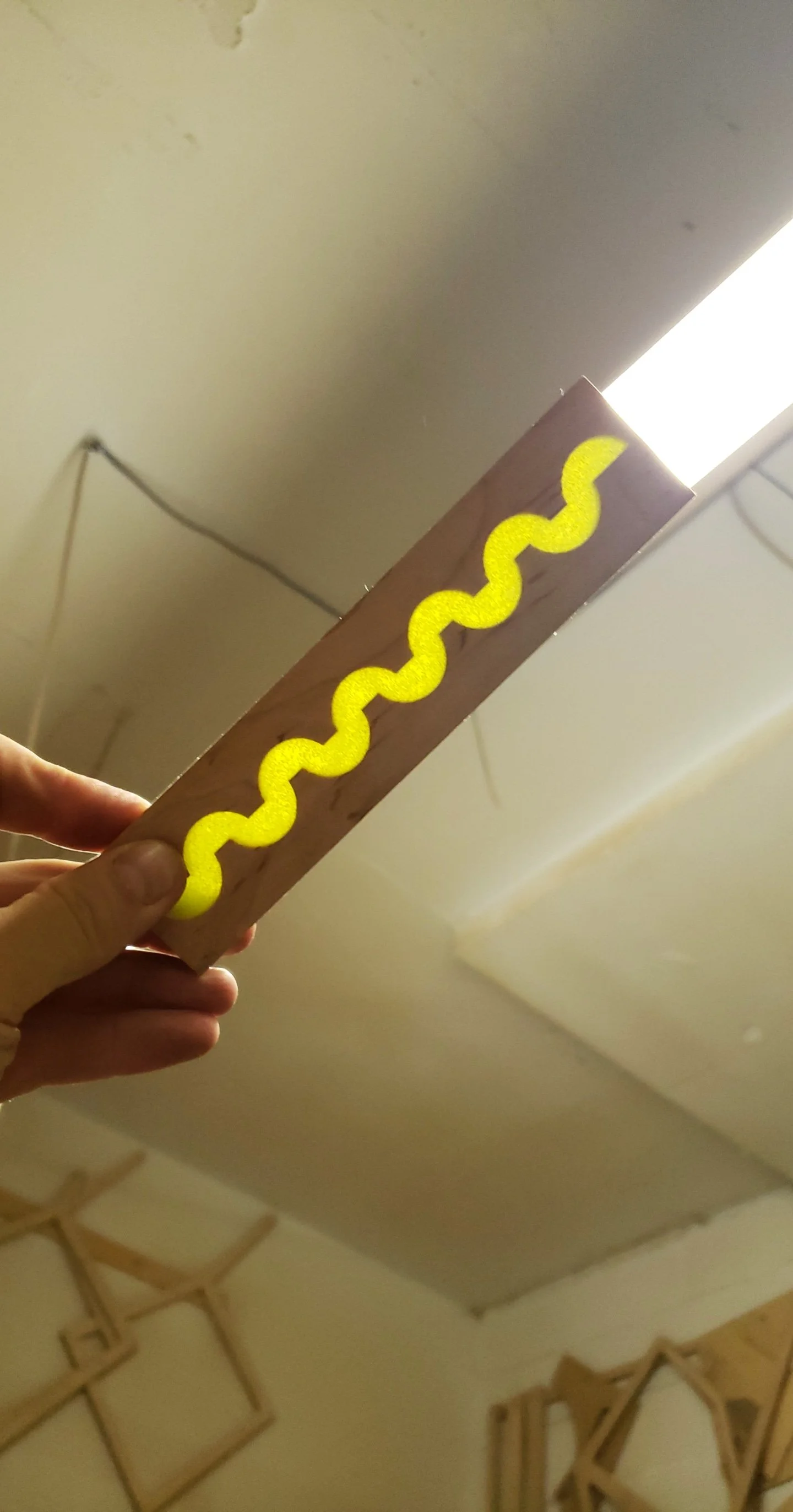 A hand holding a wooden board with a yellow, curly line painted on it, illuminated from above by a bright light.