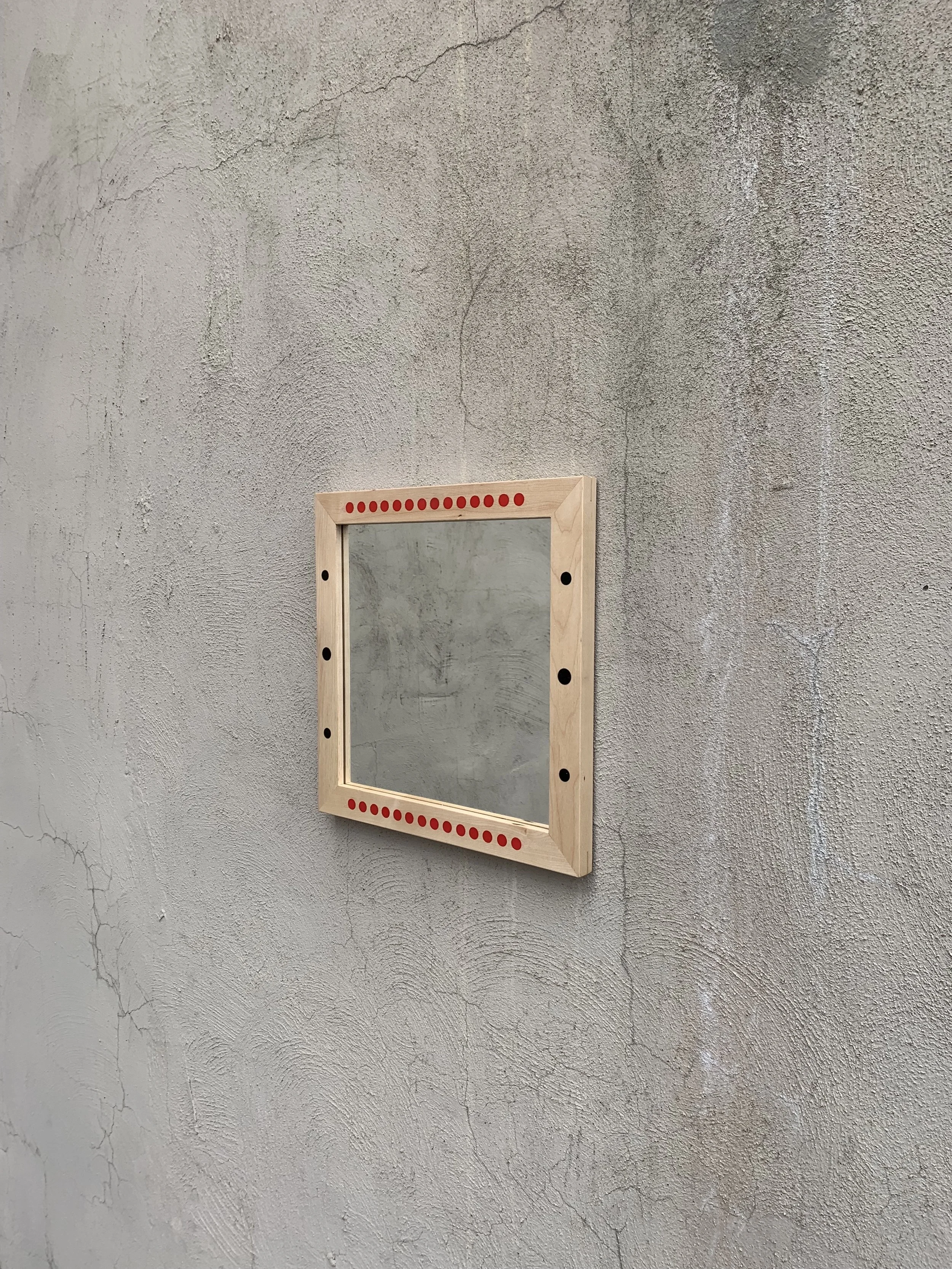 A small square mirror framed with light wood, mounted on a textured concrete wall. The frame has red and black decorative dots.