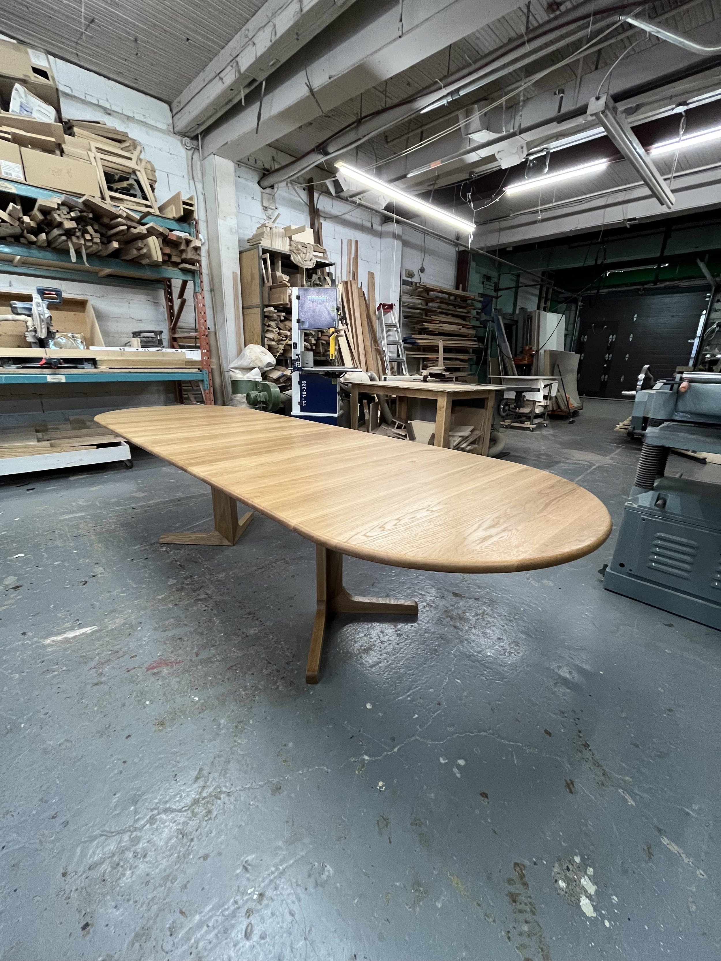 A large wooden dining table in a woodworking workshop surrounded by tools, wood planks, and shelves with woodworking materials.